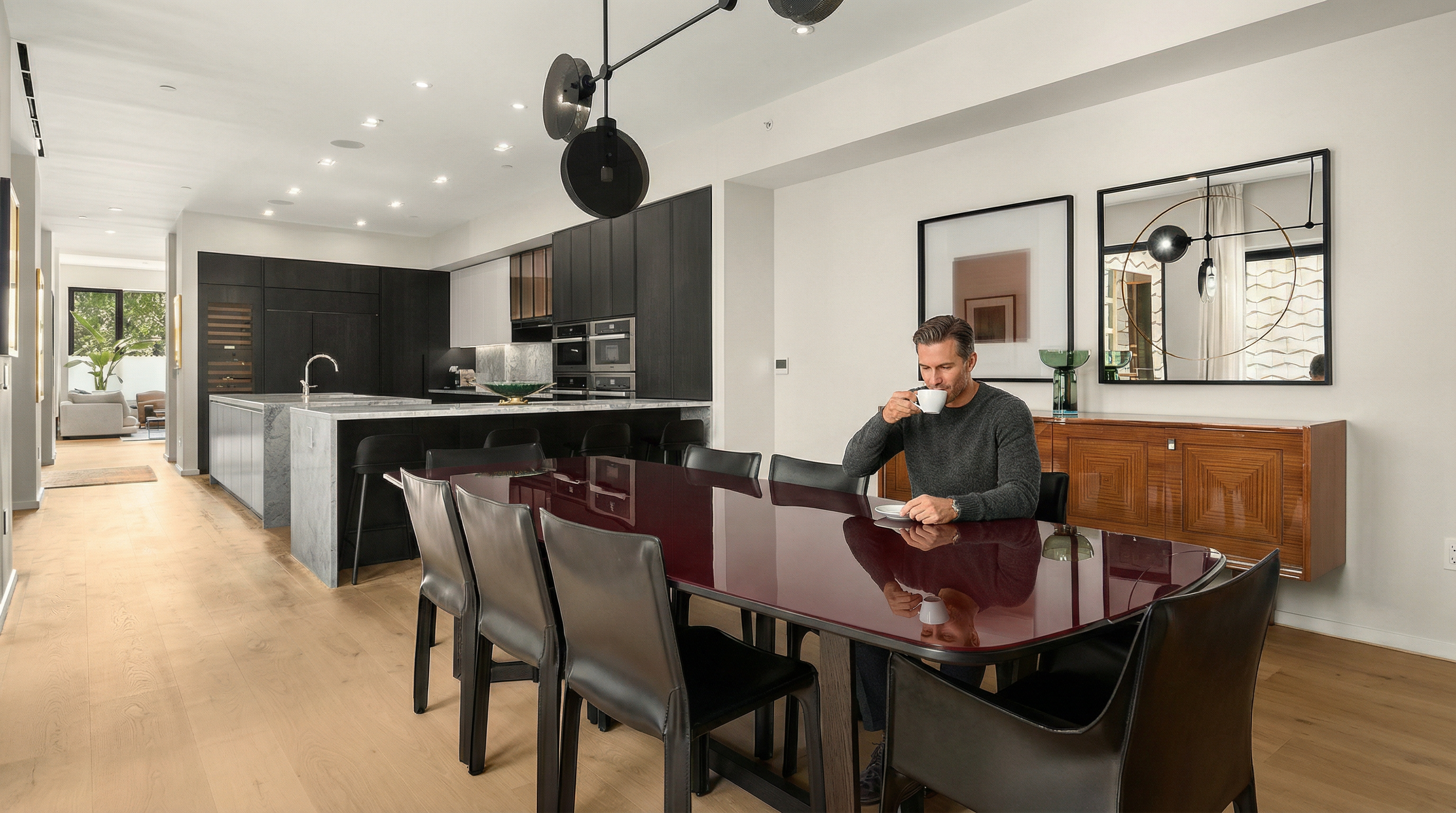 This interior shot showcases a modern dining room adjacent to a kitchen area. The dining room features a large, dark-toned dining table with matching chairs, illuminated by a contemporary chandelier. A wooden sideboard with artwork above it adds a touch of elegance, while the hardwood floors contribute to the room's warm and inviting atmosphere.