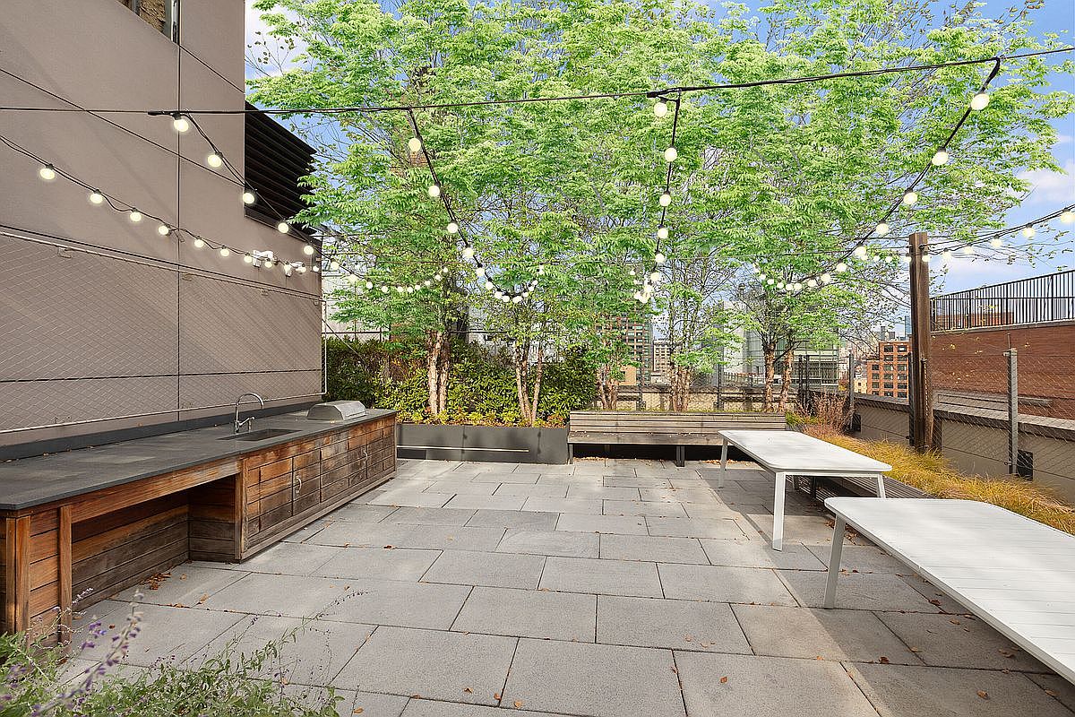This image showcases a stylish outdoor patio area, featuring a built-in wooden kitchen counter with a sink and grill, complemented by modern white tables and a wooden bench. String lights are strung above, creating a warm and inviting atmosphere, while lush greenery in the background adds a touch of nature to the urban setting. The patio is paved with large gray tiles, providing a clean and contemporary look.