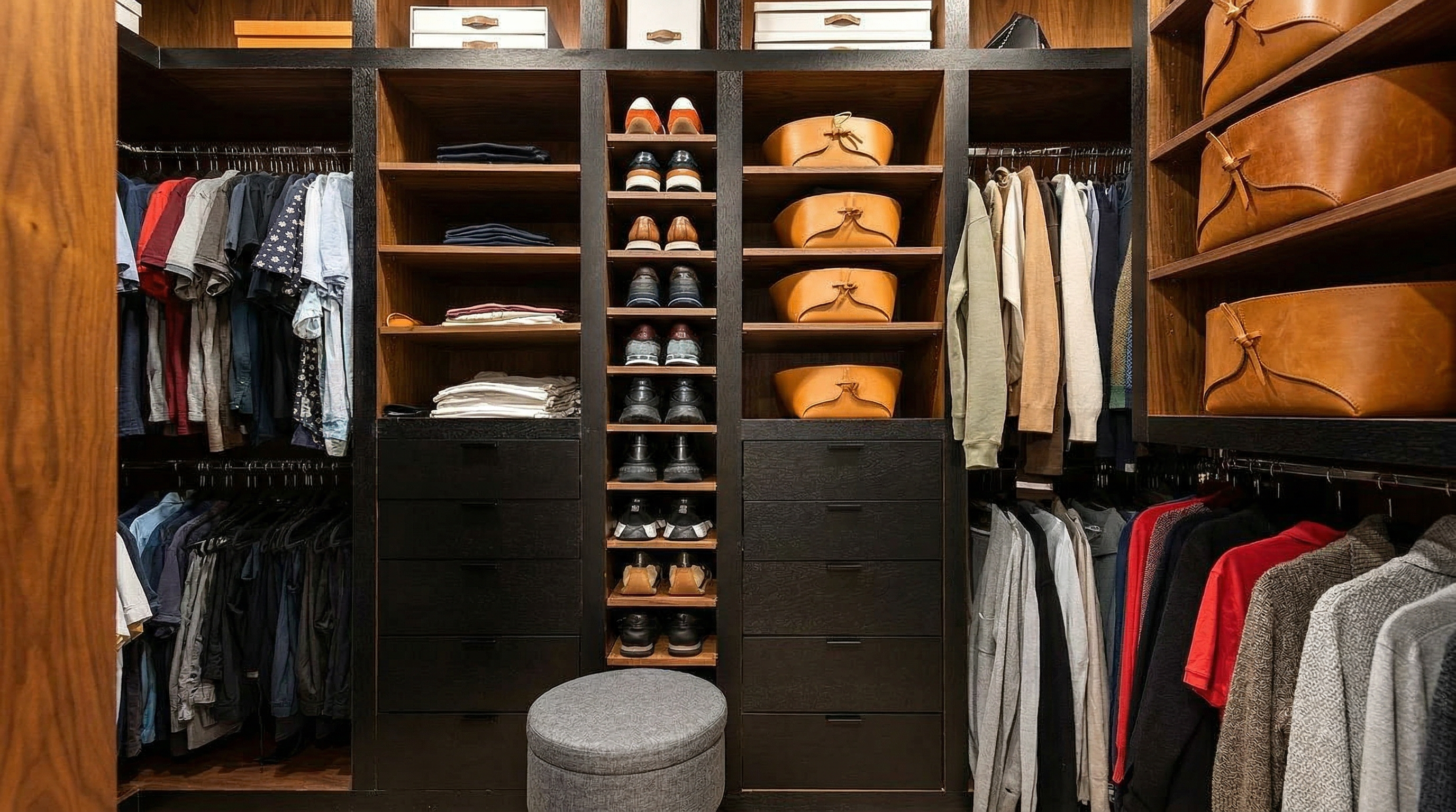 This is a well-organized walk-in closet featuring custom wood shelving and drawers. The closet is filled with clothing, shoes, and accessories, all neatly arranged. A gray stool sits in the center of the closet, providing a place to sit while getting dressed.