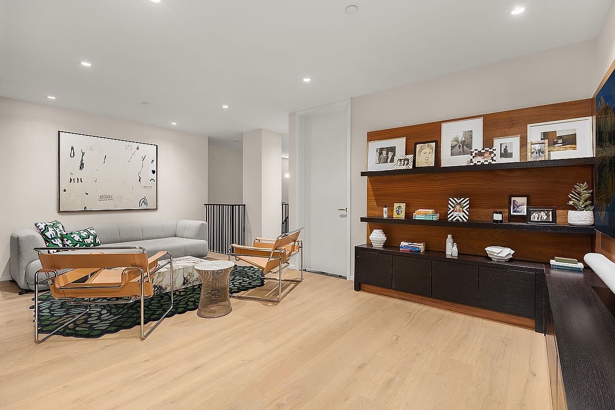 This is a modern living room featuring a gray sofa with decorative pillows, two leather and chrome chairs, and a unique coffee table. A large abstract painting hangs on the wall, and a built-in shelving unit with dark wood accents displays various decorative items. The room has light wood flooring and recessed lighting, creating a bright and inviting atmosphere.