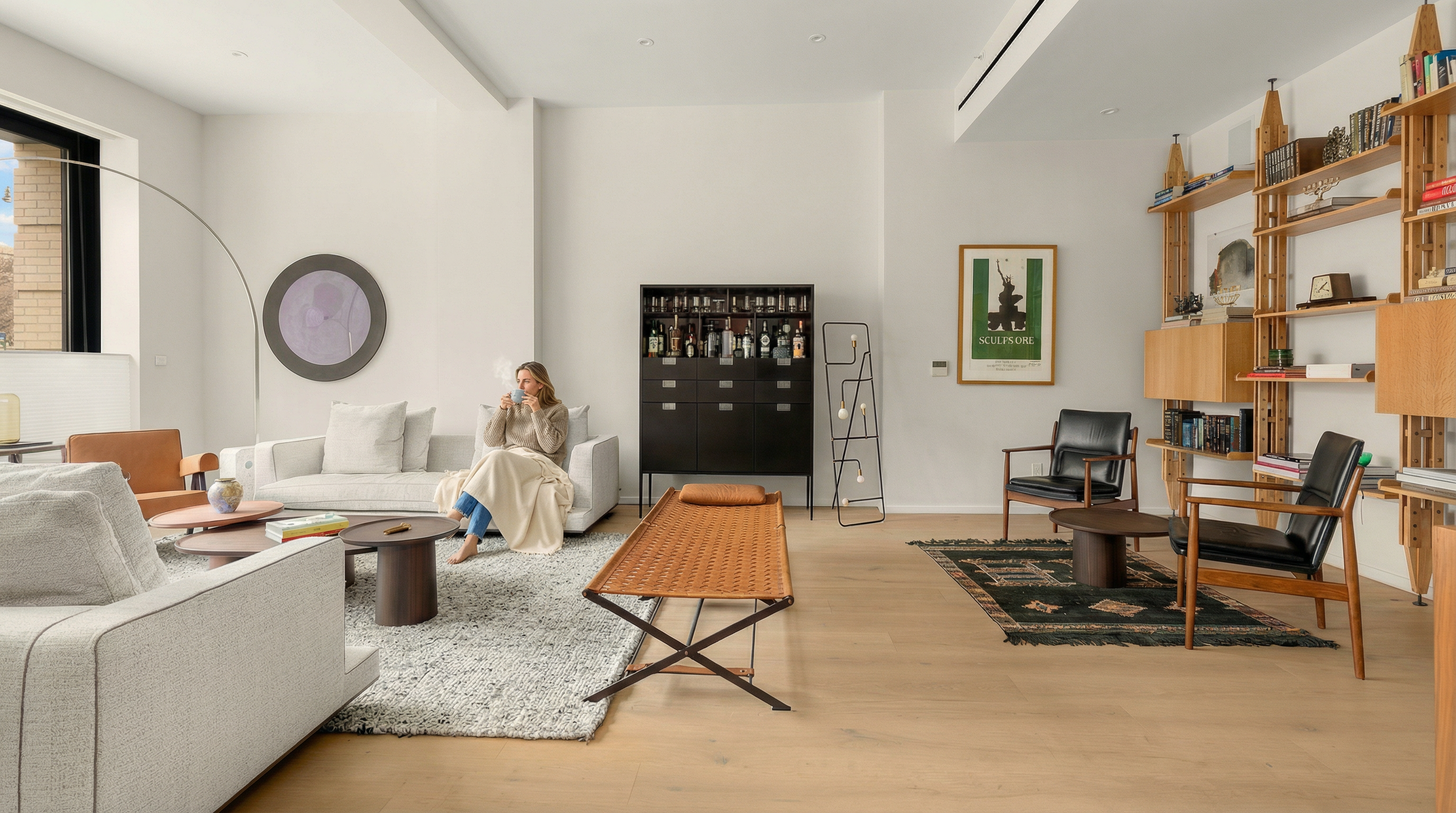 This is a spacious and well-lit living room featuring modern furniture and decor. The room includes two sofas, a woven bench, and two armchairs arranged around coffee tables and rugs. A black cabinet and built-in shelving provide storage and display space, while artwork and a floor lamp add visual interest. The hardwood flooring and neutral color palette create a sophisticated and inviting atmosphere.