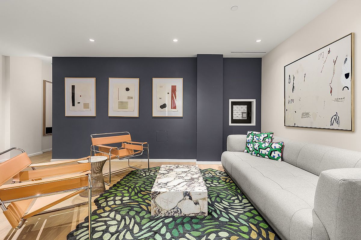 This is an interior shot of a modern living room featuring a light gray sofa with decorative pillows, a marble coffee table, and two tan leather chairs. The room is decorated with abstract art pieces on a dark blue wall, complemented by a patterned green and black rug. The overall impression is stylish and contemporary, creating a comfortable and visually appealing space.
