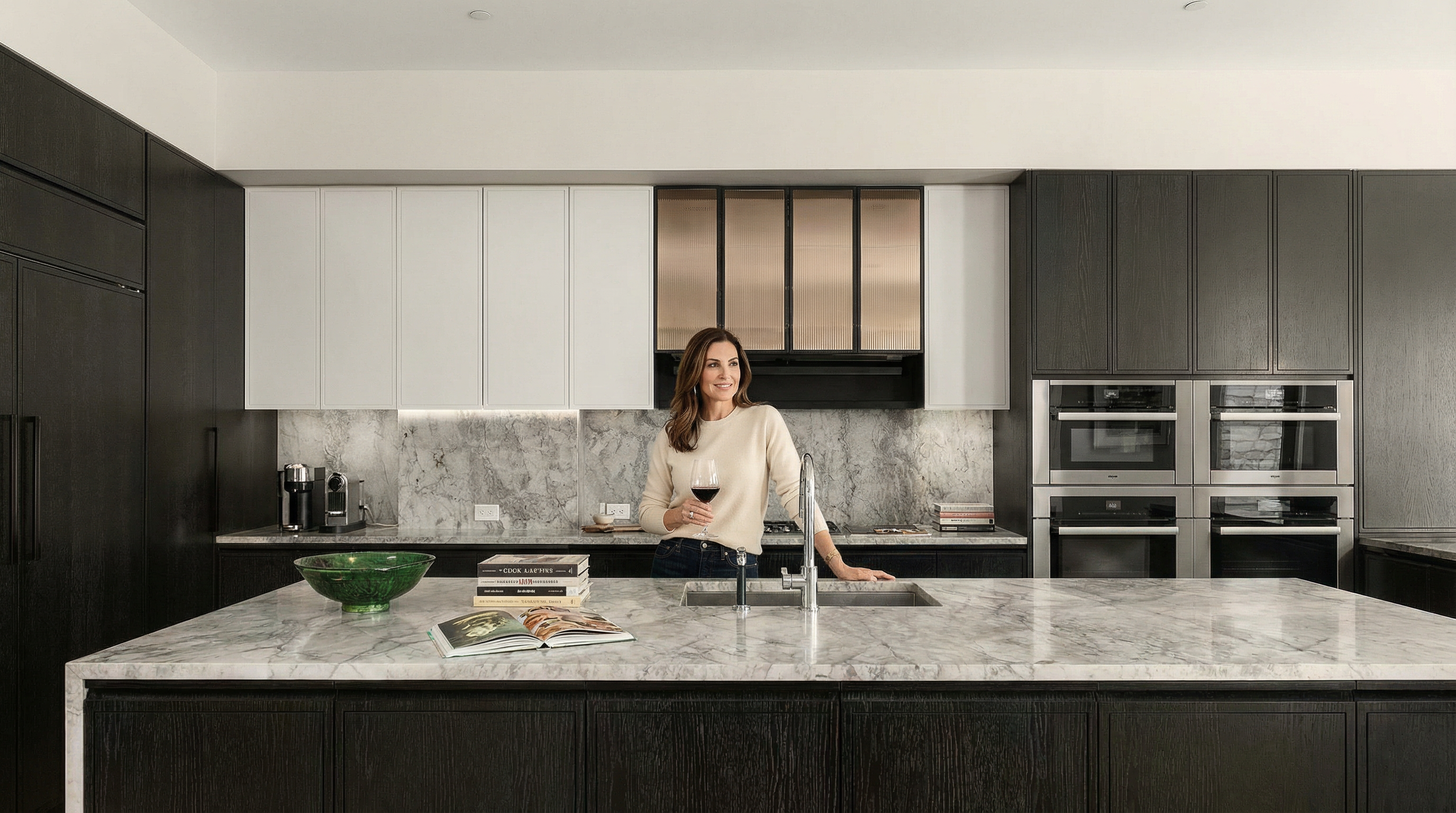 This is a modern kitchen with dark wood cabinetry and a large island with a marble countertop. The kitchen features stainless steel appliances and a marble backsplash. The perspective is from the front of the island, showcasing the kitchen's layout and design.