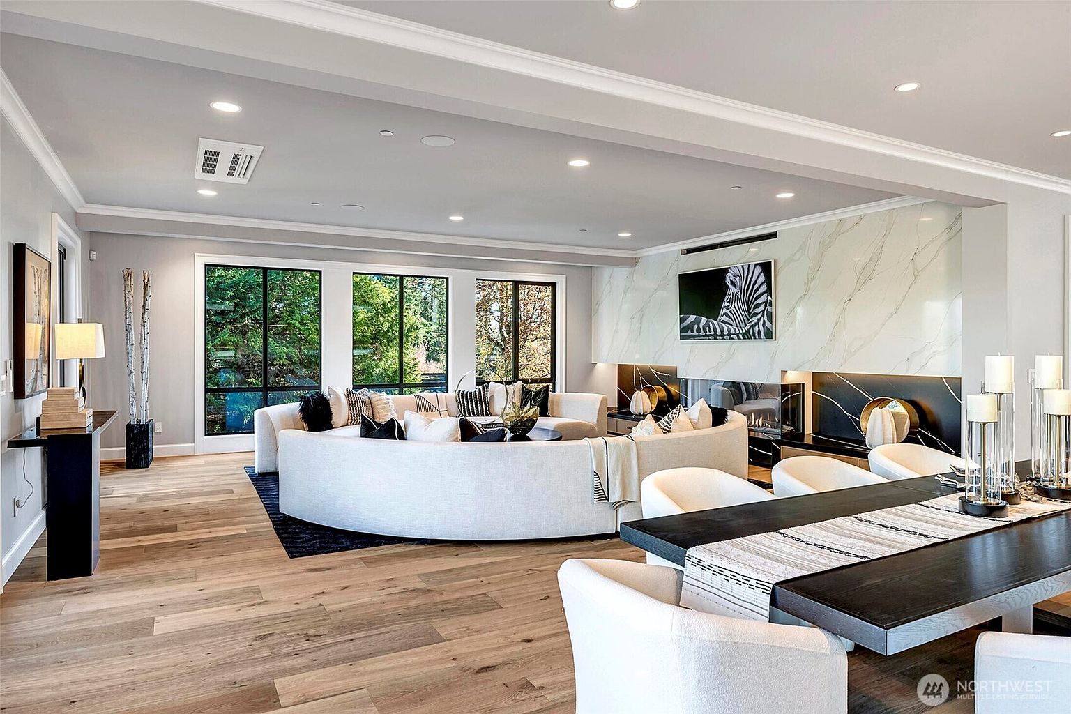 This expansive, open-concept living room features a sophisticated modern aesthetic with light hardwood flooring and a dramatic, curved white sofa as the central focal point. A striking floor-to-ceiling marble fireplace wall with integrated black accents and a zebra-themed art piece anchors the space, while large windows provide abundant natural light and views of the surrounding greenery. The room is elegantly furnished with a dining area in the foreground, creating a seamless flow perfect for entertaining.