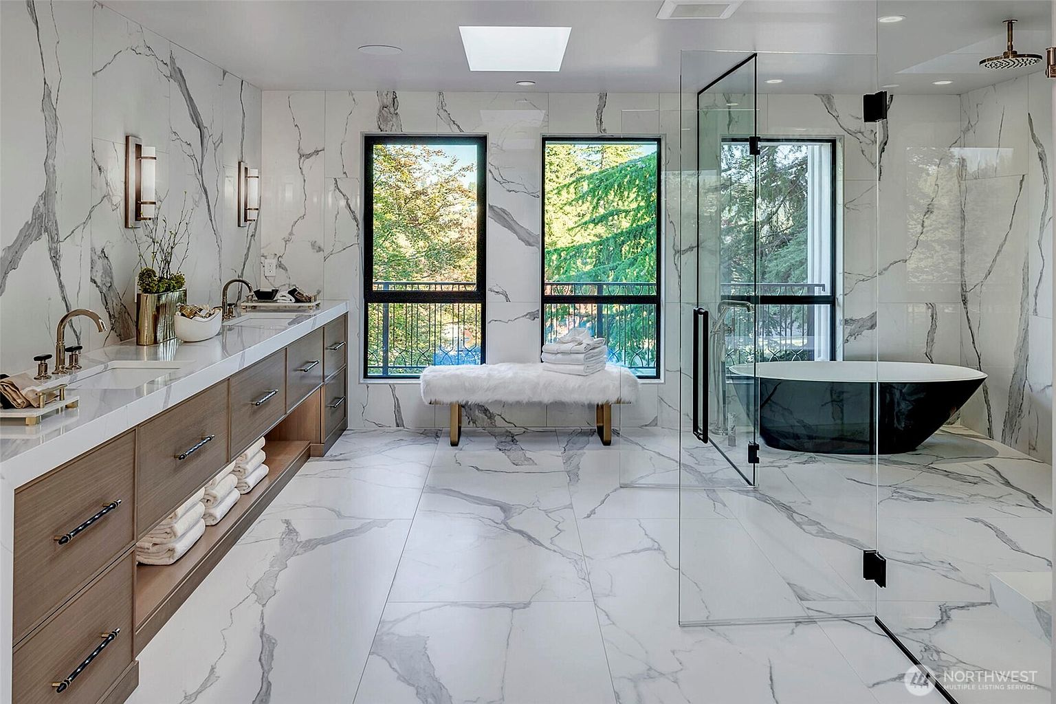 This luxurious primary bathroom features floor-to-ceiling marble-look tile, a sleek double vanity with wood cabinetry, and a spacious glass-enclosed walk-in shower. A freestanding black soaking tub sits elegantly by the window, while a plush bench and skylight add to the spa-like atmosphere. The perspective is a wide-angle shot that captures the sophisticated, high-end design and bright, airy feel of the space.