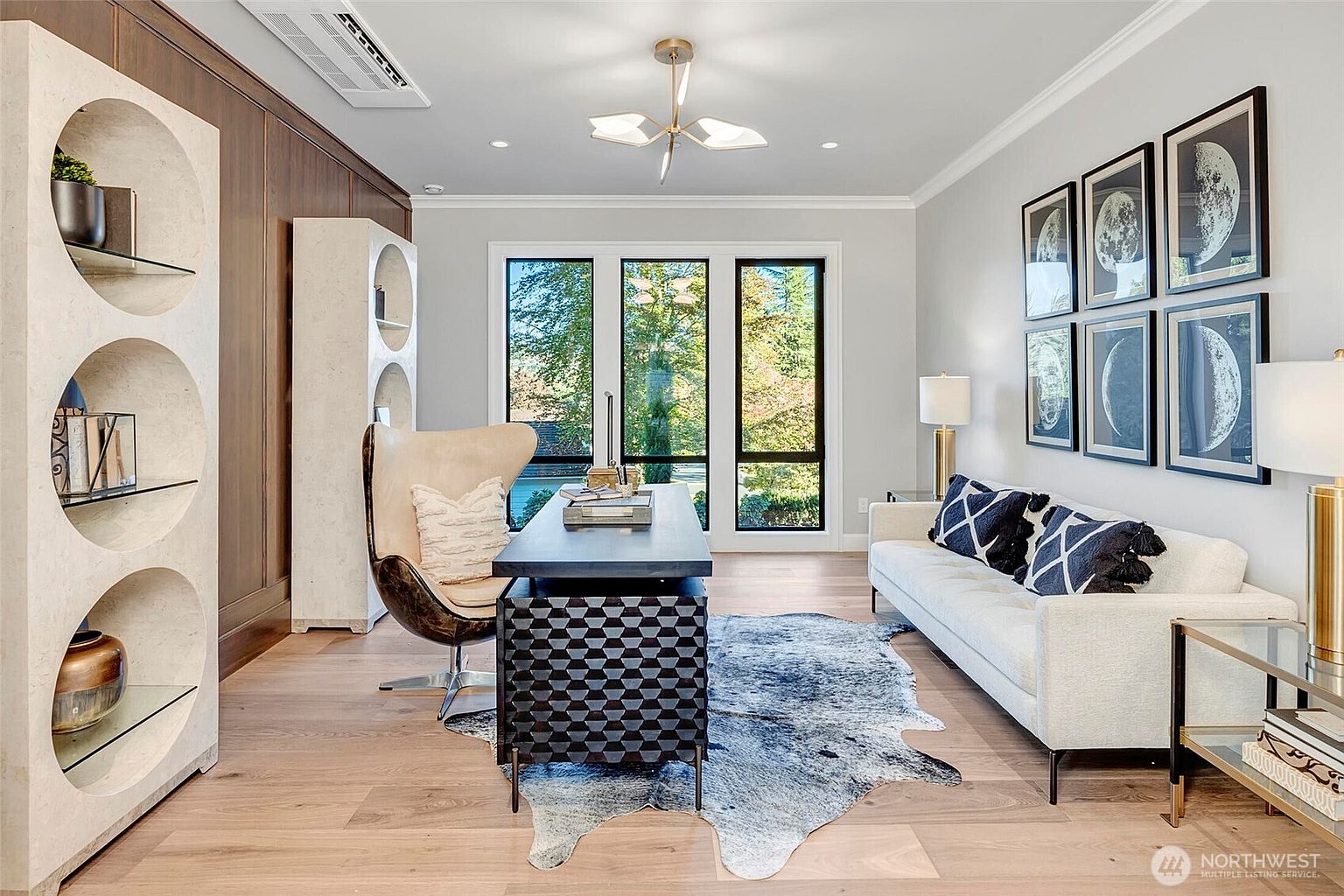 This sophisticated home office features a striking geometric-patterned desk paired with a modern cream-colored swivel chair, set against a backdrop of floor-to-ceiling wood paneling and a unique arched shelving unit. A plush, cowhide-style area rug anchors the space, while a sleek white sofa adorned with patterned pillows provides a comfortable seating area beneath a gallery wall of moon-themed art. The room is bathed in natural light from large windows, creating a professional yet inviting atmosphere perfect for productivity.