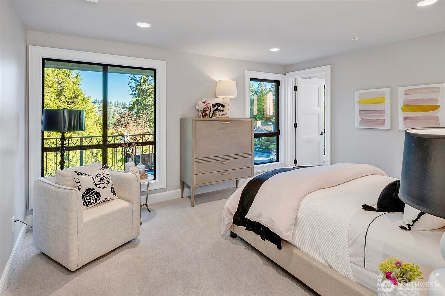 This bright and airy primary bedroom features a neutral color palette, plush carpeting, and a comfortable armchair positioned near a large window with a scenic outdoor view. A sleek, light-wood dresser sits between two windows, while a neatly made bed with layered bedding serves as the room's focal point. The space is accented by modern wall art and elegant lighting, creating a serene and sophisticated atmosphere.