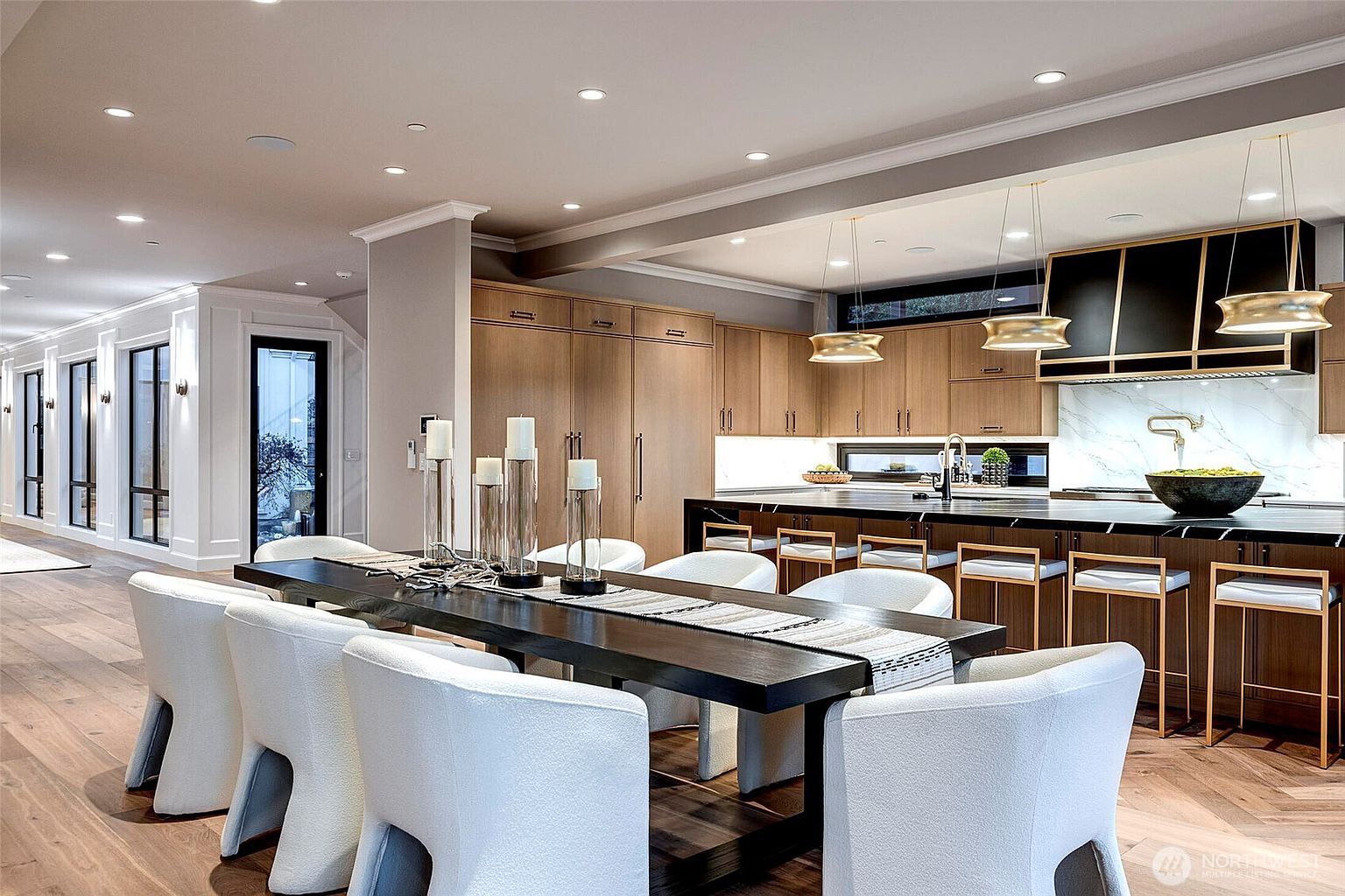 This sophisticated open-concept kitchen and dining area features a long, dark wood dining table paired with modern white upholstered chairs, leading into a sleek kitchen with light wood cabinetry and a large black marble-topped island. The space is illuminated by elegant gold pendant lights and recessed lighting, creating a warm and luxurious atmosphere. The perspective captures the seamless flow between the dining and culinary zones, highlighting the high-end finishes and contemporary design.