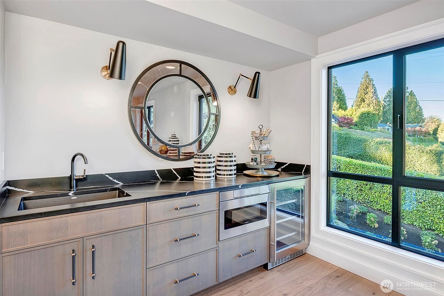 This modern wet bar area features sleek, light-toned wood cabinetry paired with a striking black marble countertop and integrated sink. A large, circular decorative mirror hangs above the counter, flanked by two contemporary wall sconces, while a built-in microwave and wine refrigerator add functional luxury. The space is bathed in natural light from a large window that offers a serene view of the lush, manicured greenery outside.