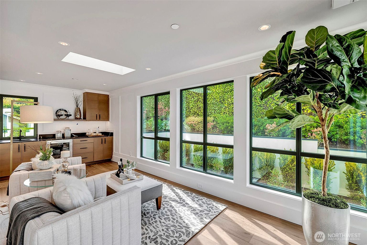 This bright and airy living room features a contemporary design with light-toned wood flooring, a plush grey sofa, and a patterned area rug. Large floor-to-ceiling windows flood the space with natural light and provide a serene view of the lush green garden outside, while a sleek kitchenette area with wooden cabinetry is tucked into the corner. The room is accented by a large potted fiddle leaf fig, creating a sophisticated and tranquil atmosphere perfect for relaxation.