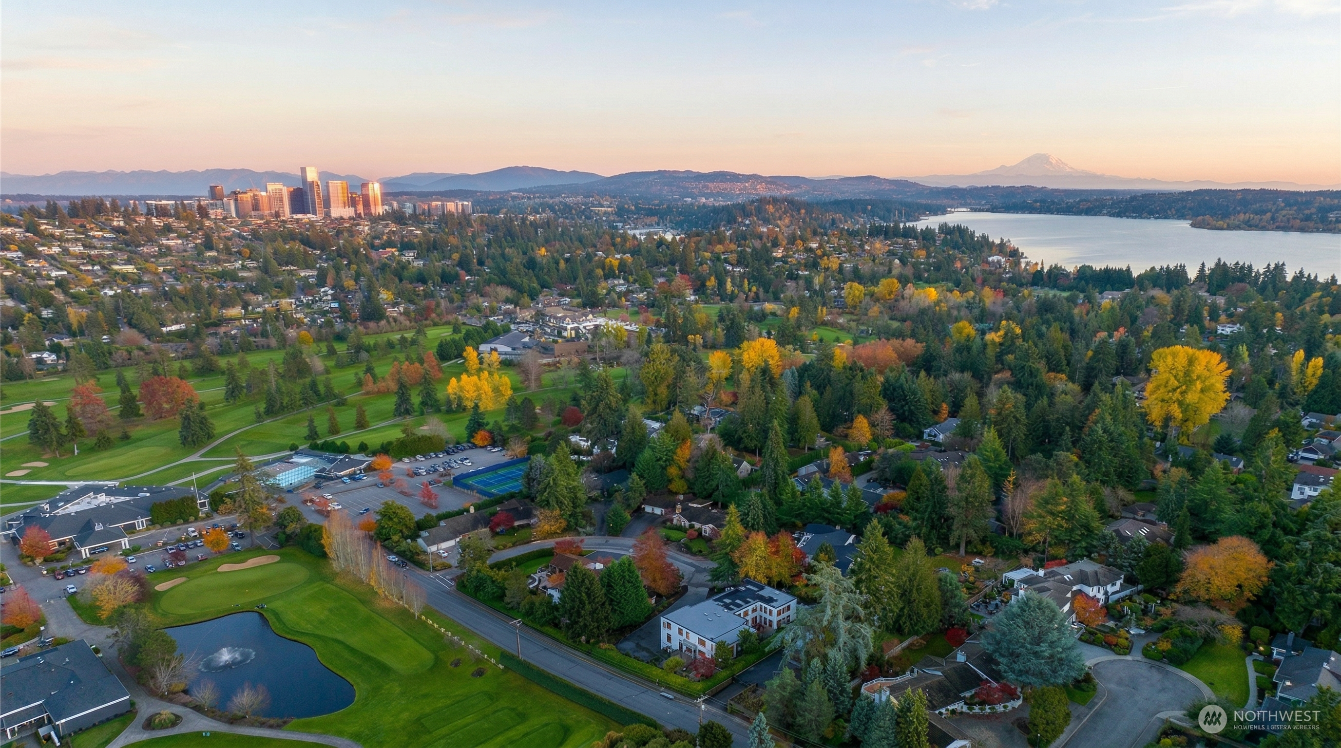 This high-angle aerial view captures a residential property nestled within a lush, tree-lined neighborhood adjacent to a golf course. The perspective showcases the home's location relative to a scenic lake, the city skyline in the distance, and the prominent silhouette of Mount Rainier on the horizon. An arrow points directly to the subject property, highlighting its integration into the serene, park-like surroundings.