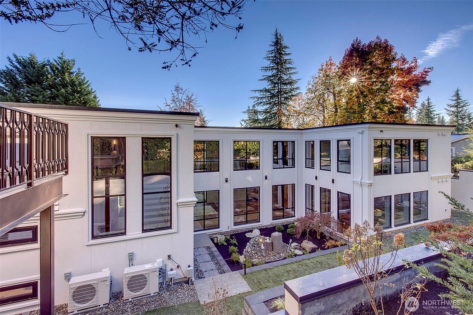 This striking modern home features a clean, white stucco facade with expansive, multi-pane black-framed windows that create a sophisticated, geometric aesthetic. The rear of the house wraps around a beautifully landscaped courtyard garden, complete with a stone water feature and manicured plantings. The perspective is elevated, capturing the architectural harmony between the contemporary structure and the surrounding mature trees under a clear blue sky.
