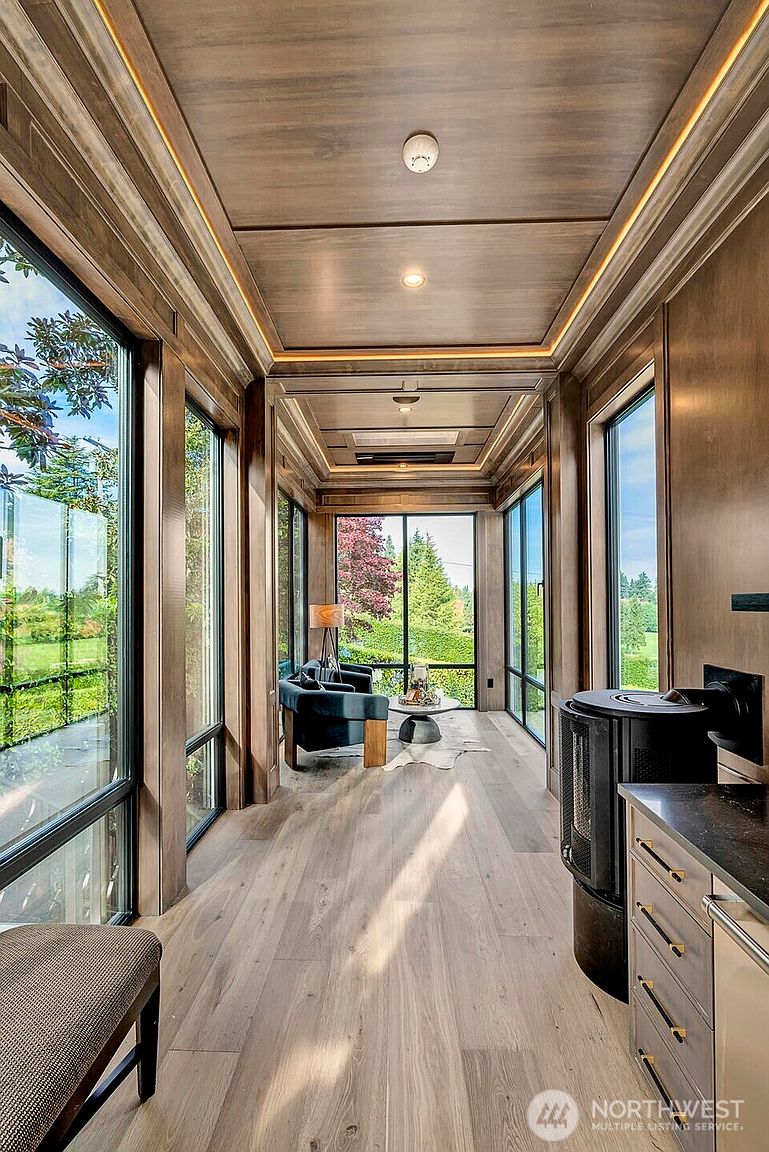 This elegant sunroom features floor-to-ceiling windows that flood the space with natural light and offer expansive views of the surrounding greenery. The room is finished with rich, warm wood paneling on the walls and ceiling, complemented by light-toned hardwood flooring and a cozy seating area at the far end. A modern, cylindrical wood-burning stove adds a sophisticated touch to this serene and inviting retreat.