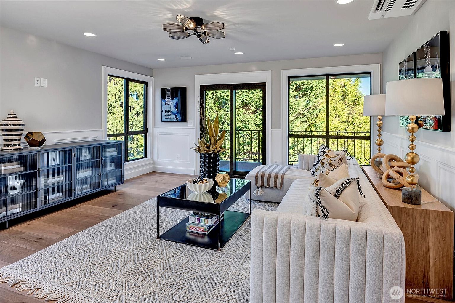 This sophisticated living room features a modern, neutral-toned aesthetic with a plush, ribbed-texture sofa and a sleek black-framed glass coffee table. The space is anchored by a large, patterned area rug and a striking dark blue media console, complemented by elegant gold-accented table lamps and a unique geometric ceiling fixture. Large windows and a sliding glass door provide ample natural light and a view of the lush greenery outside, creating an inviting and upscale atmosphere.