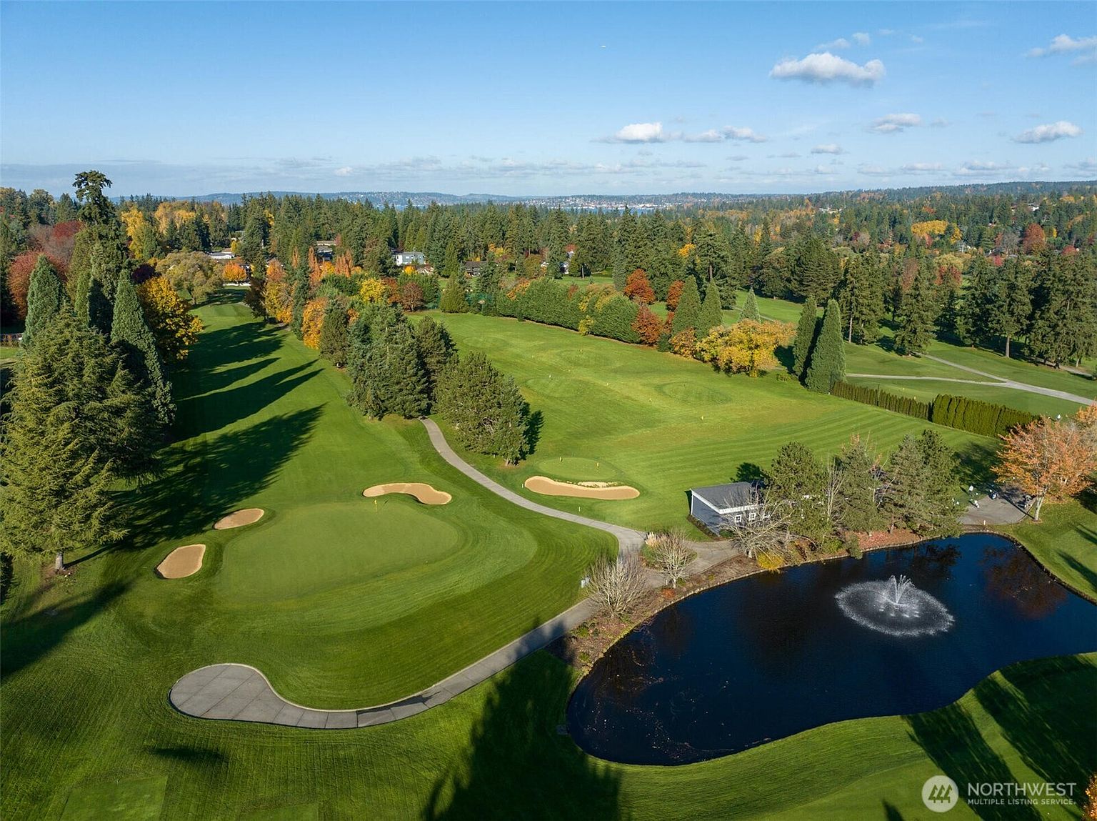 This high-angle aerial view captures a lush, well-manicured golf course featuring a vibrant green fairway, sand bunkers, and a serene pond with a fountain. The landscape is framed by dense, mature trees displaying autumn colors, creating a peaceful and expansive recreational setting. The perspective emphasizes the community's access to premium outdoor amenities and scenic natural beauty.