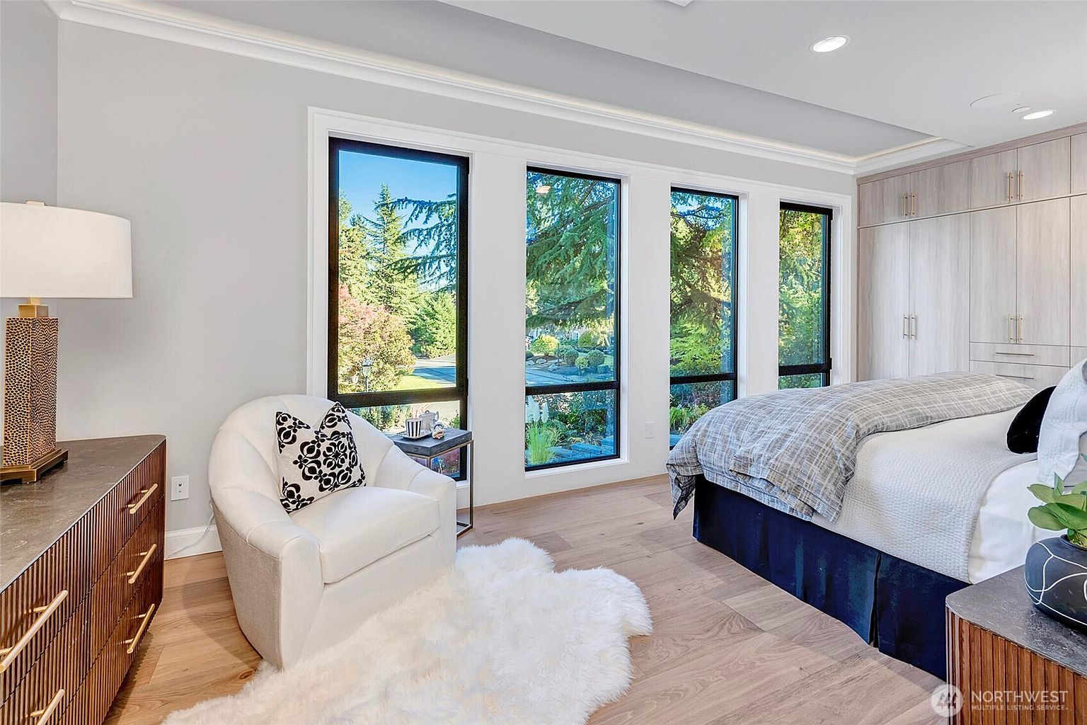 This elegant primary bedroom features light hardwood flooring, a plush white armchair with a faux fur rug, and a bed dressed in a sophisticated plaid duvet. Large, floor-to-ceiling windows flood the room with natural light and offer a serene view of the surrounding greenery, while built-in cabinetry provides ample storage. The space exudes a calm, contemporary luxury with a neutral color palette and high-end finishes.