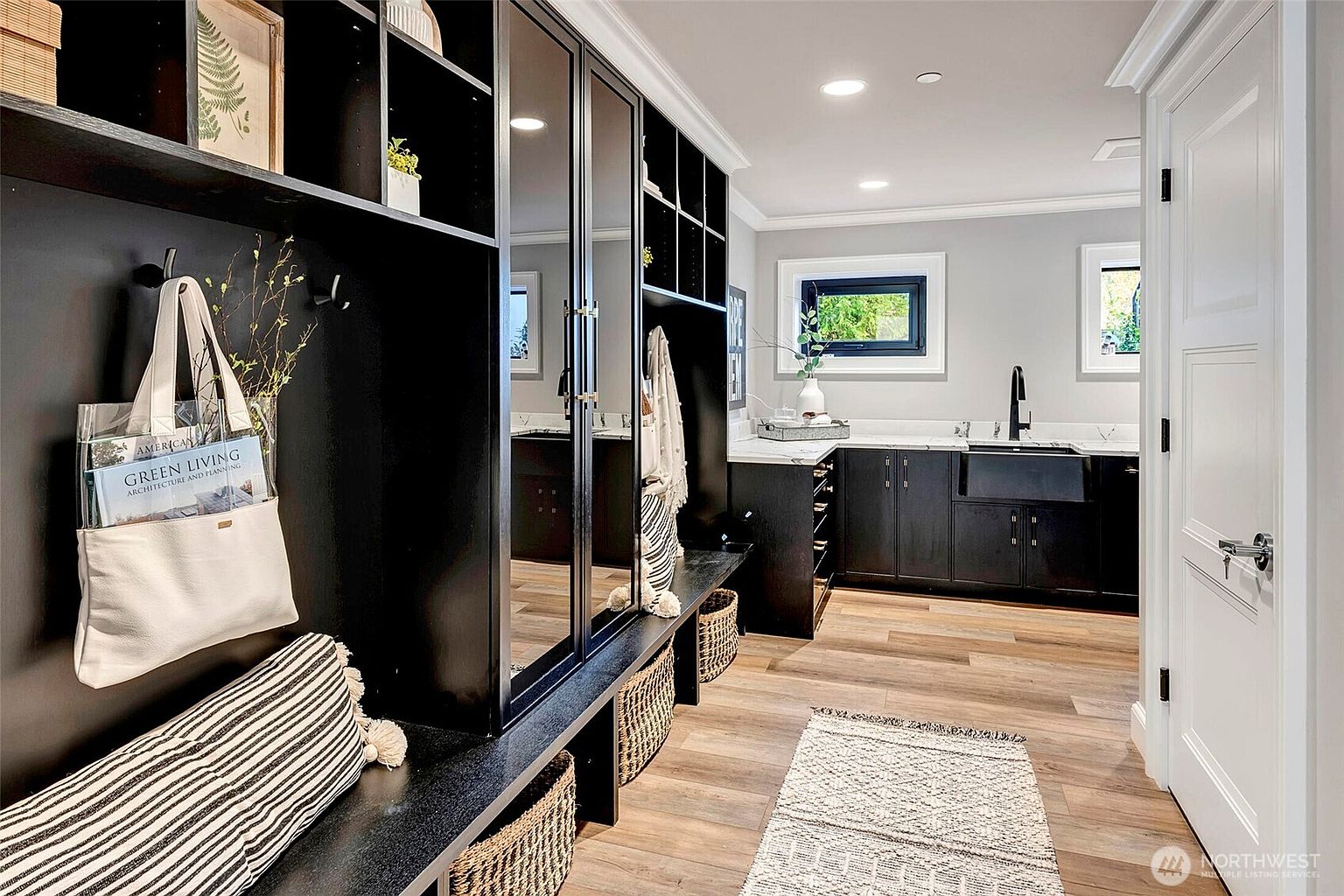 This sophisticated mudroom and laundry area features sleek black cabinetry, a farmhouse sink, and warm wood-look flooring. The space is highly functional, incorporating built-in storage cubbies, a bench with striped cushions, and a mirror-fronted closet, creating a modern and organized aesthetic. The perspective captures the transition from the entryway storage into the utility area, highlighting the clean lines and high-end finishes.