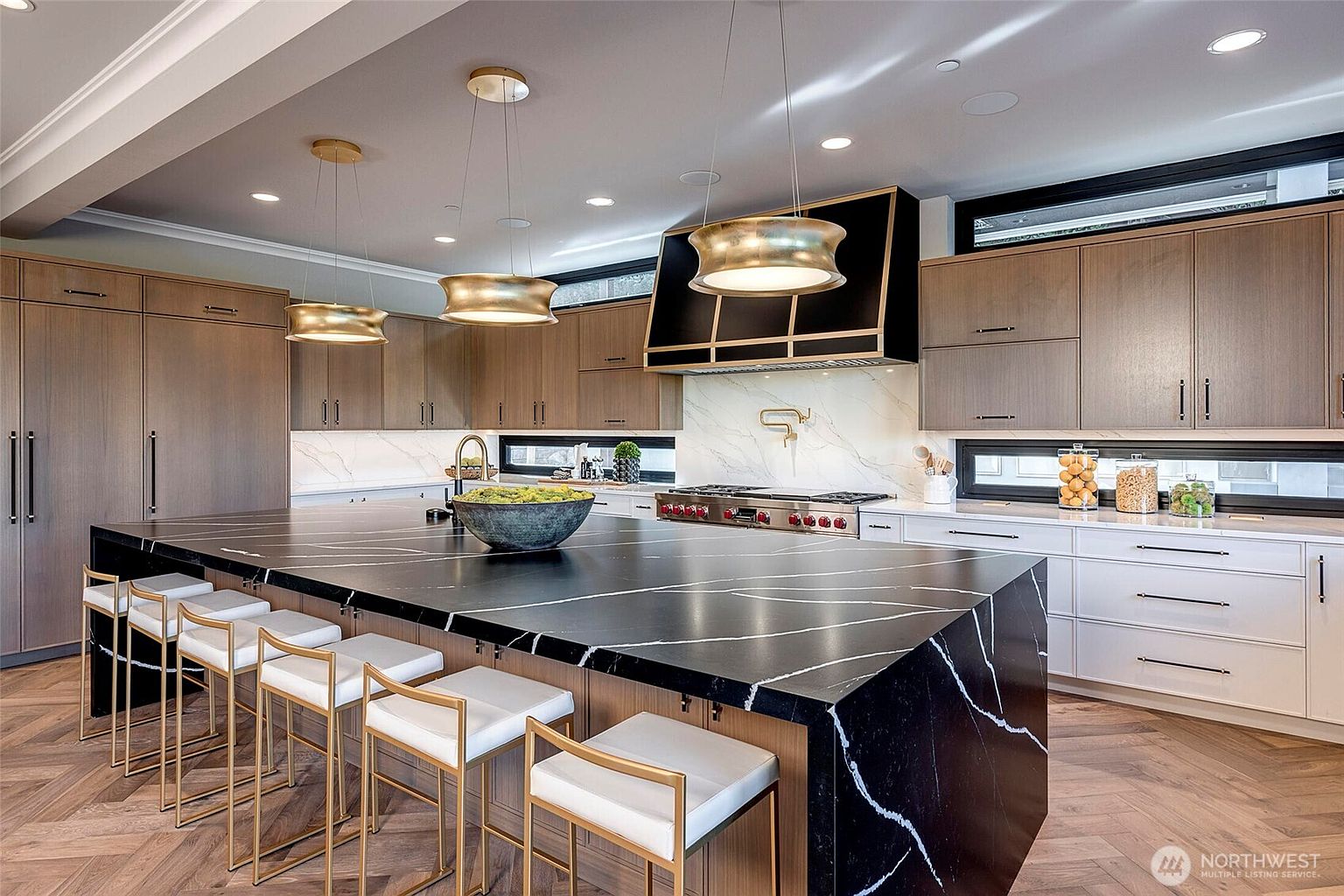 This sophisticated, modern kitchen features a striking black marble waterfall island with white veining, complemented by warm wood-toned cabinetry and crisp white lower cabinets. The space is anchored by a bold, black-and-gold range hood and elegant gold pendant lighting, all set against a herringbone-patterned hardwood floor. The perspective is a wide-angle shot from the kitchen island, showcasing a high-end, luxurious design perfect for both cooking and entertaining.