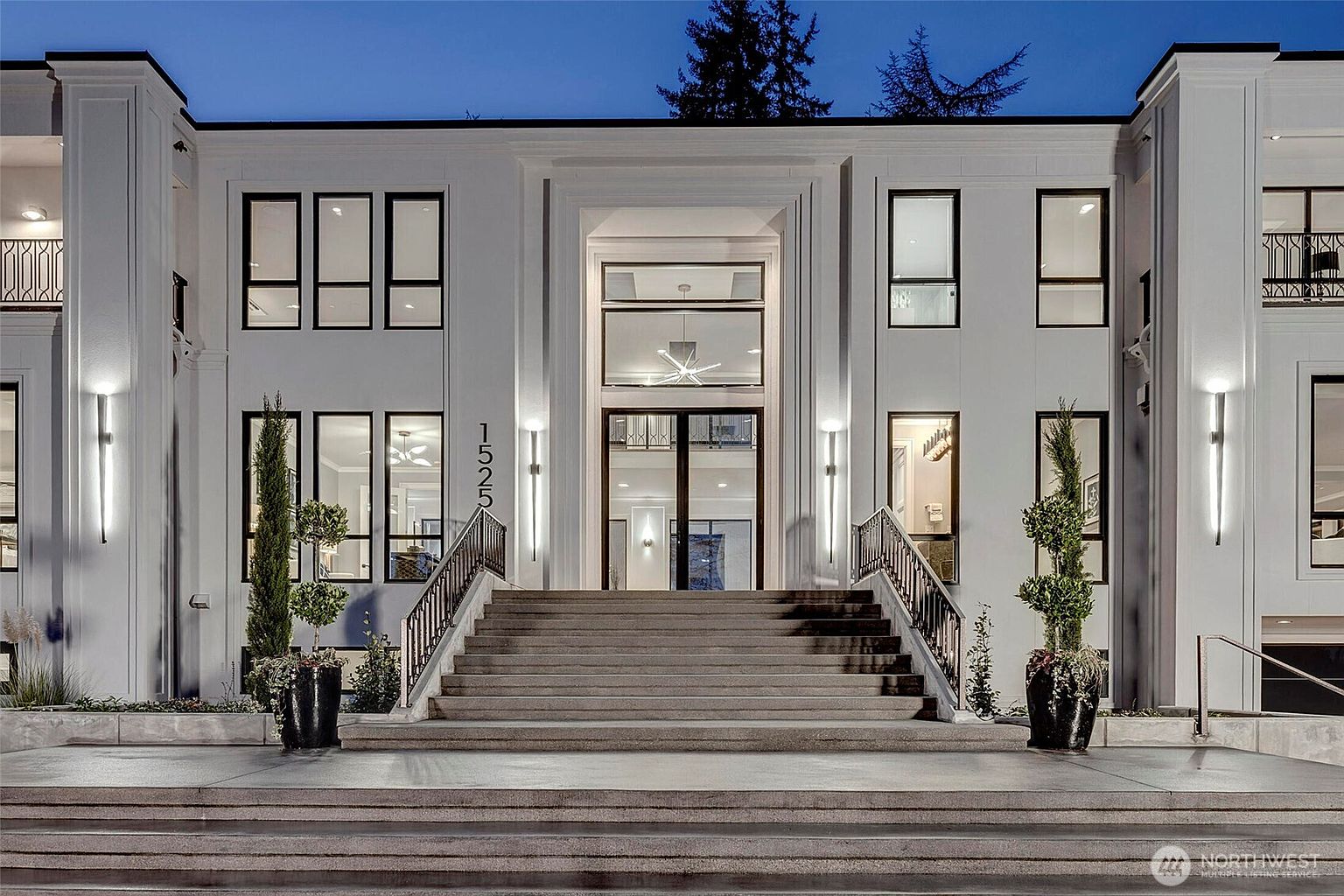 This grand, modern estate features a symmetrical white facade with clean lines and a prominent central entryway accessed by a wide, tiered stone staircase. The exterior is illuminated by sleek, vertical wall-mounted lights, highlighting the architectural details and the manicured topiary plants flanking the entrance. The perspective is a low-angle, eye-level shot that emphasizes the imposing scale and sophisticated, contemporary design of the residence at twilight.