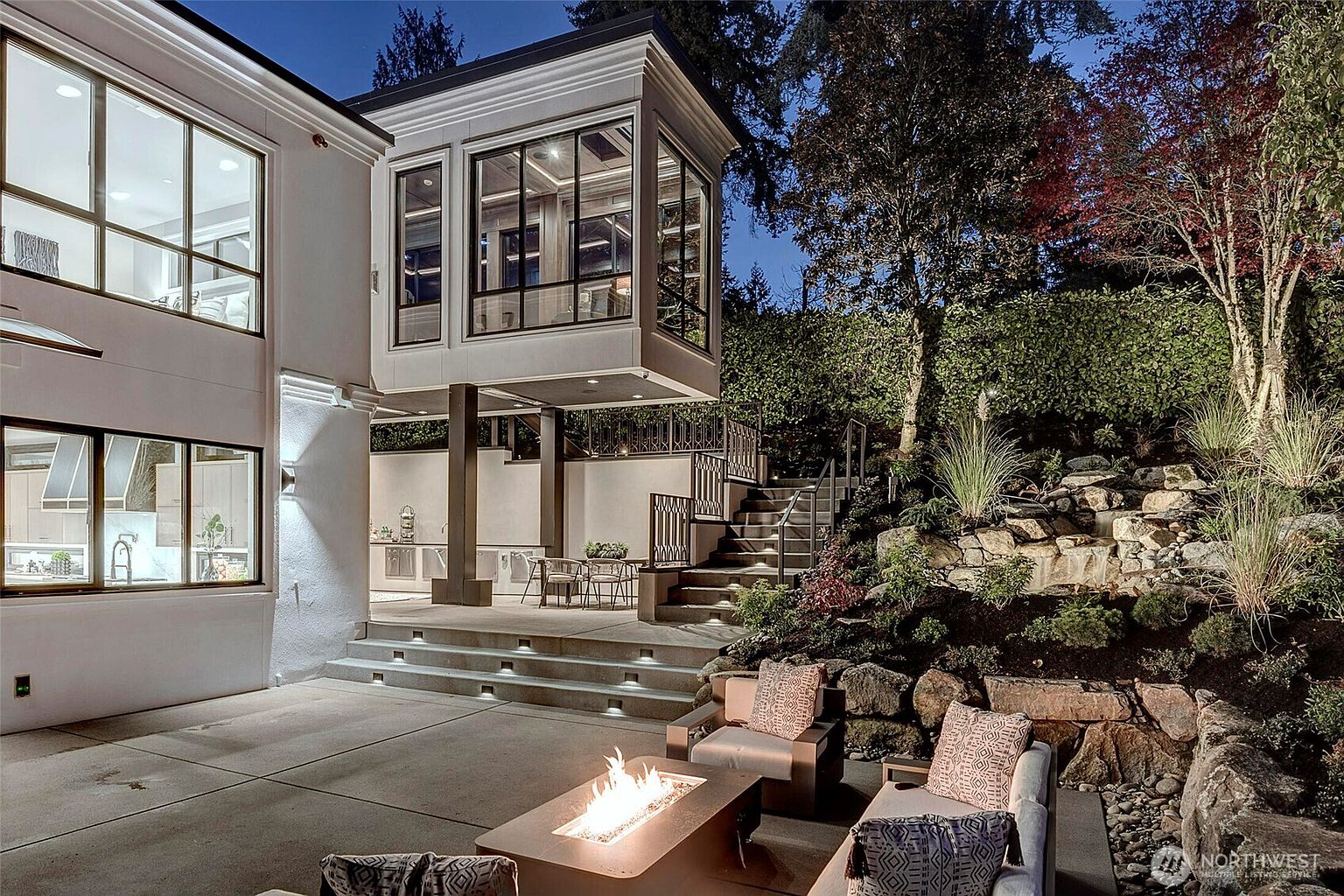 This sophisticated outdoor living space features a modern concrete patio with a built-in fire pit and comfortable lounge seating, perfect for evening entertaining. The area is framed by a striking architectural cantilevered room above, a stone-retaining wall with integrated landscaping, and a set of stairs leading to an upper terrace. The scene is captured at dusk, highlighting the warm glow of the fire and the subtle landscape lighting that creates an inviting, luxurious atmosphere.