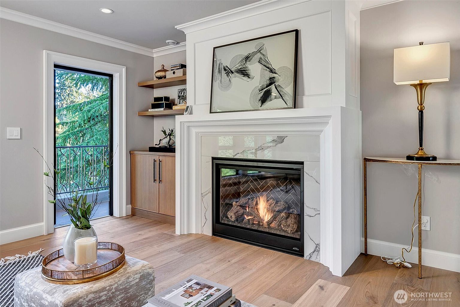 This elegant living room features a prominent white fireplace surround with marble tiling and a herringbone-patterned firebox, serving as the room's focal point. To the left, built-in wooden cabinetry and floating shelves provide sophisticated storage, while a glass door leads out to a balcony with a view of lush greenery. The space is finished with light hardwood floors, a neutral color palette, and a stylish table lamp, creating a warm and inviting atmosphere.