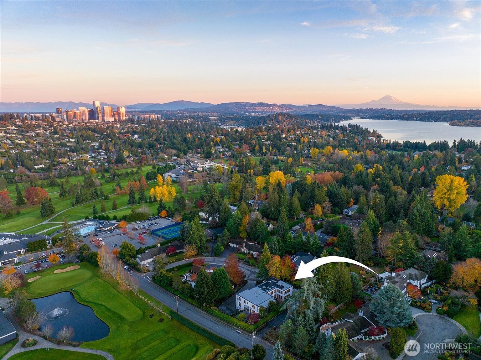 This high-angle aerial view captures a residential property nestled within a lush, tree-lined neighborhood adjacent to a golf course. The perspective showcases the home's location relative to a scenic lake, the city skyline in the distance, and the prominent silhouette of Mount Rainier on the horizon. An arrow points directly to the subject property, highlighting its integration into the serene, park-like surroundings.
