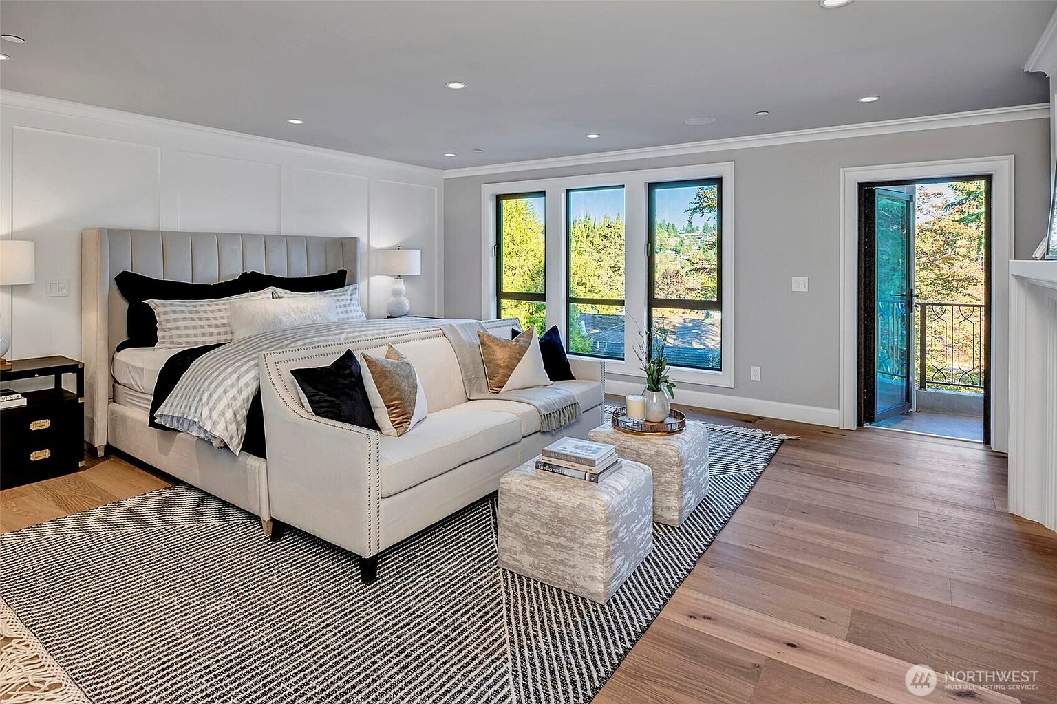 This elegant primary bedroom features a plush, tufted headboard bed paired with a sophisticated sofa at its foot, creating a luxurious lounge area. The room is anchored by a large, patterned area rug over light hardwood floors, with floor-to-ceiling windows that flood the space with natural light and offer a view of the balcony. The neutral color palette, combined with recessed lighting and clean architectural lines, provides a serene and high-end atmosphere.