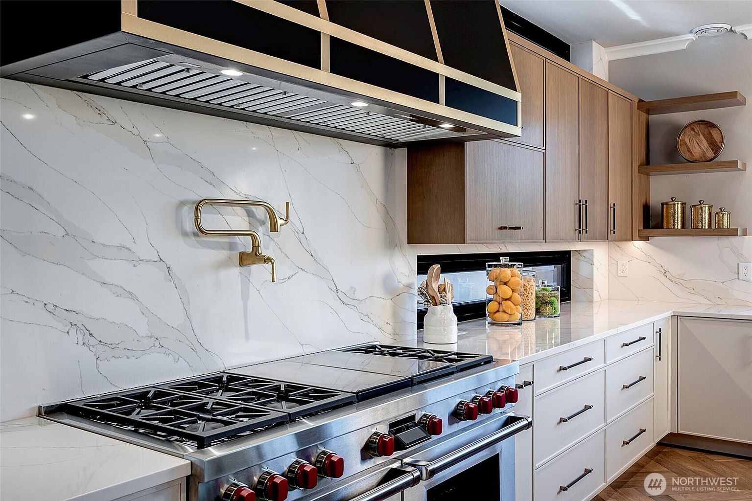 This high-end kitchen features a professional-grade stainless steel range with distinctive red knobs, set against a striking white marble-veined backsplash. A custom black and gold range hood serves as a bold focal point, complemented by warm wood cabinetry and open shelving. The space is elegantly designed with a pot filler faucet and a sleek, modern aesthetic that emphasizes luxury and functionality.