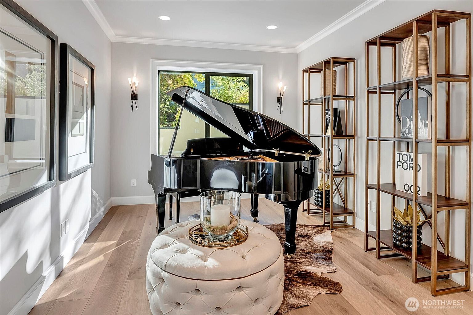 This elegant music room or study features a polished black grand piano as its centerpiece, positioned on a textured cowhide rug. The room is framed by light hardwood floors, a tufted cream ottoman, and tall, open-concept gold-toned shelving units displaying curated decor. Symmetrical wall sconces and large framed art pieces add a sophisticated, refined aesthetic to the space.