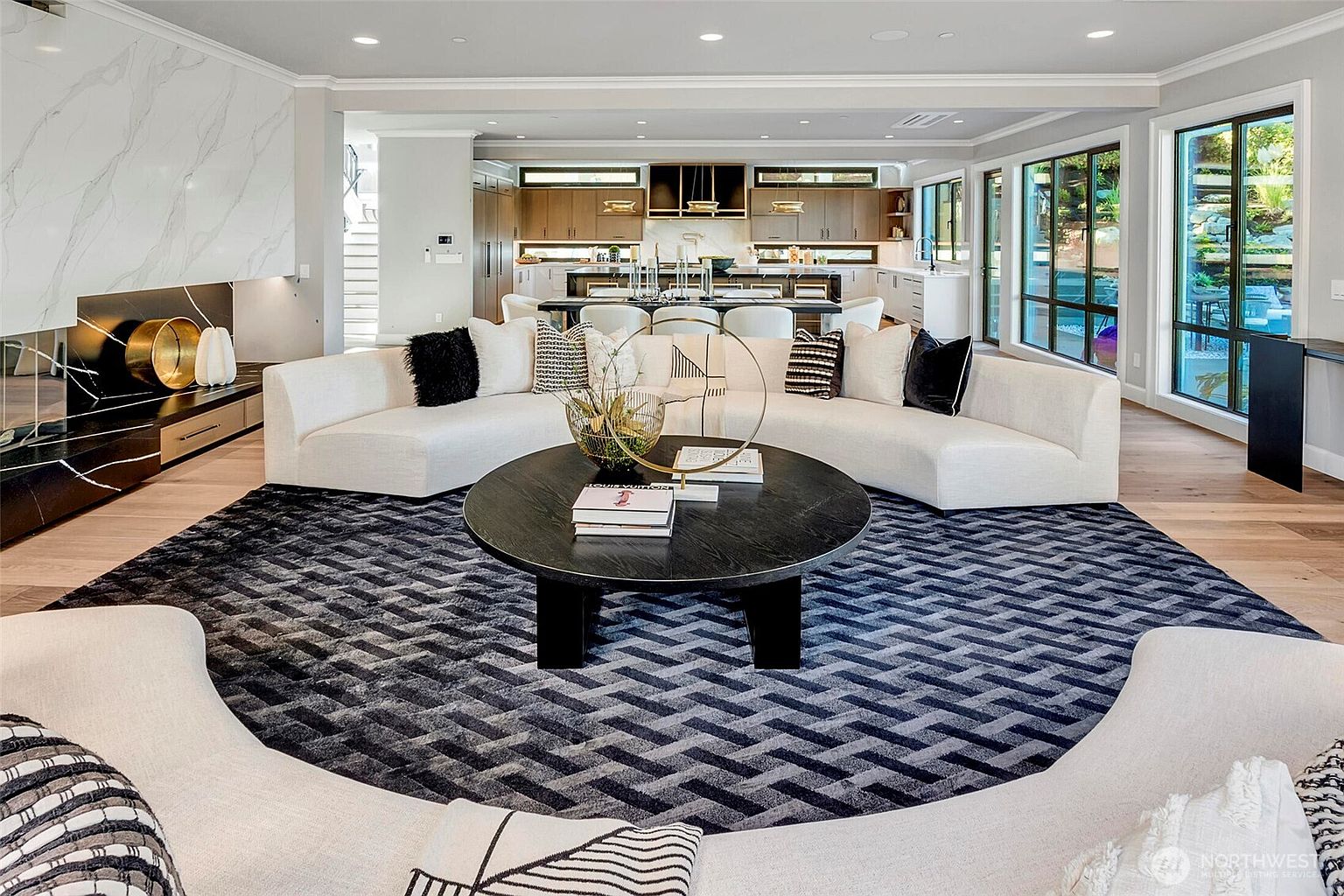 This expansive, open-concept living area features a sophisticated modern design with a large, curved white sectional sofa centered around a circular black coffee table on a textured geometric rug. To the left, a striking marble-clad fireplace adds a luxurious focal point, while the background reveals a seamless transition into a bright, high-end kitchen and dining space. The room is bathed in natural light from floor-to-ceiling windows, creating an airy, elegant atmosphere perfect for high-end entertaining.
