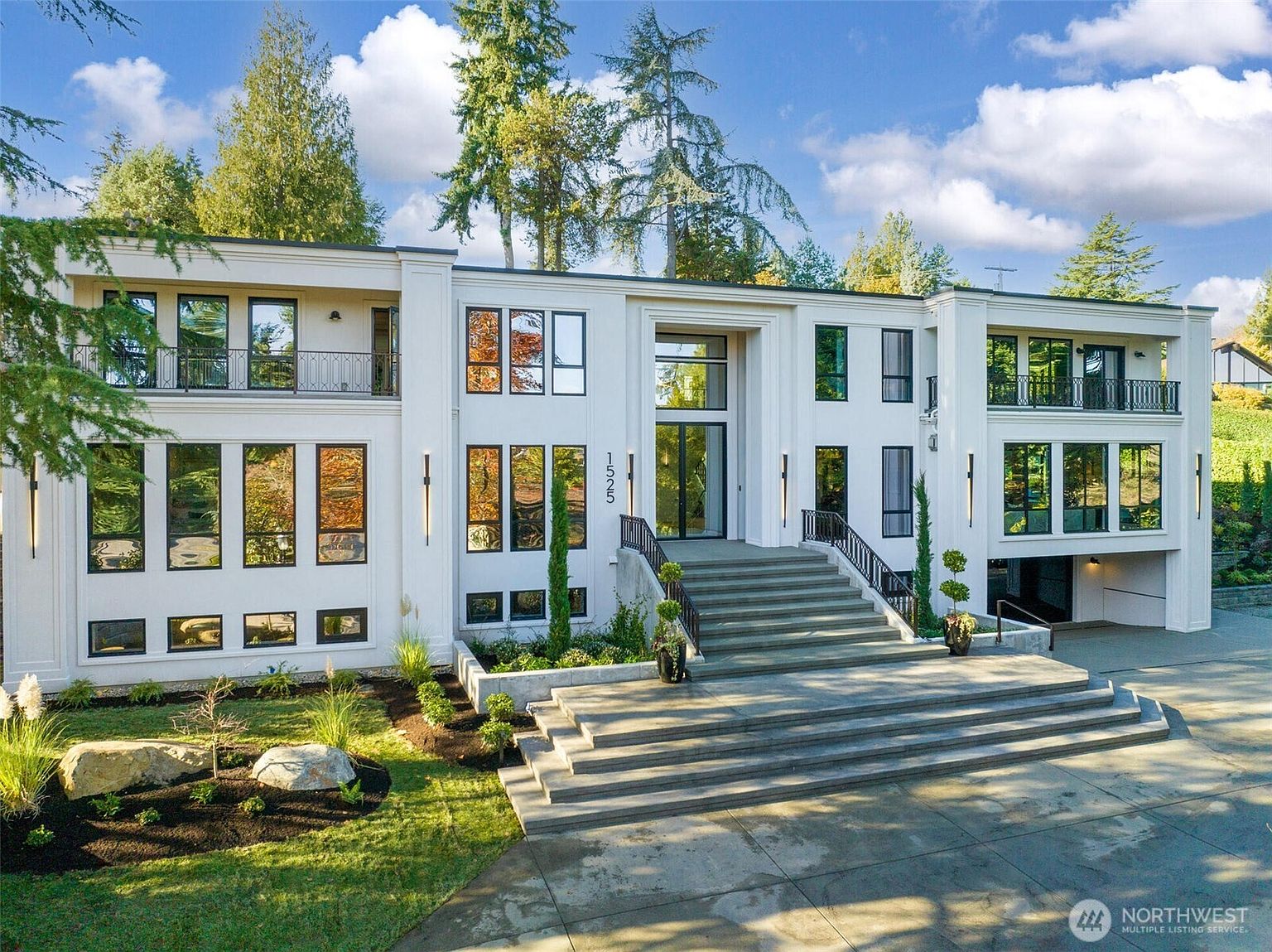 This grand, contemporary estate features a striking white facade with symmetrical architectural lines and expansive floor-to-ceiling windows. A prominent central staircase leads up to the main entrance, flanked by manicured landscaping and a paved driveway. The overall impression is one of luxury, modern elegance, and sophisticated curb appeal.