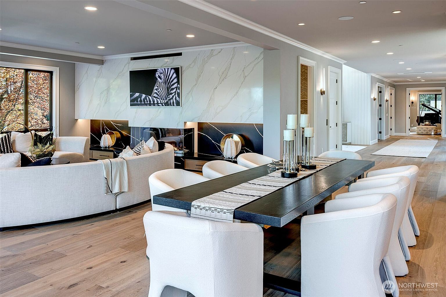 This sophisticated open-concept living and dining area features a striking floor-to-ceiling marble fireplace wall with a zebra art piece as a focal point. The space is anchored by a large, curved cream-colored sofa and a long, dark wood dining table, all set against light-toned hardwood flooring. The perspective captures a seamless flow between the cozy lounge area and the formal dining space, leading the eye toward a bright hallway in the distance.