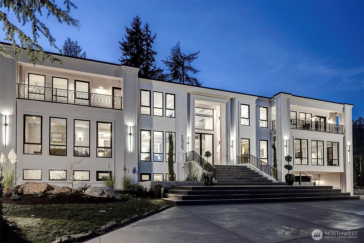 This striking, modern luxury estate is captured at dusk, showcasing its symmetrical white facade and grand, multi-tiered staircase leading to a prominent central entryway. The home features expansive floor-to-ceiling windows, elegant wrought-iron balcony railings, and sophisticated exterior lighting that highlights its architectural geometry. The perspective is a wide-angle, eye-level shot that emphasizes the imposing scale and refined curb appeal of the property.