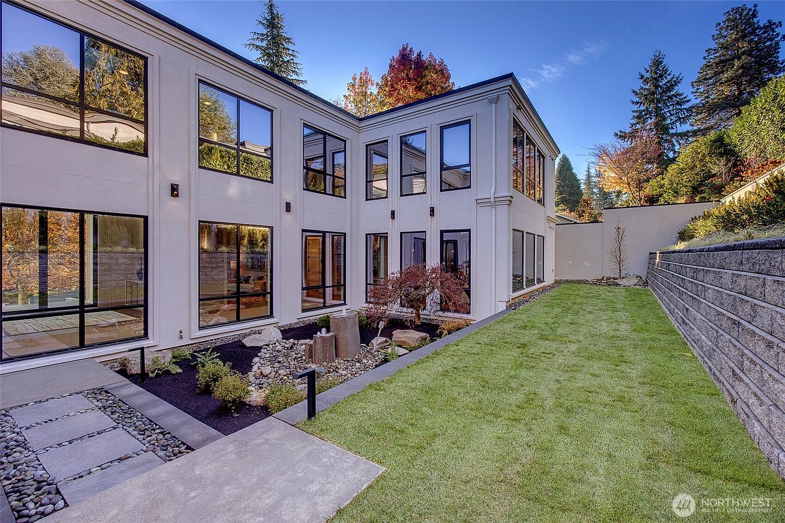 This modern, two-story home features a sleek, white stucco exterior with expansive black-framed windows that create a striking contrast. The rear yard includes a meticulously landscaped garden bed with a stone water feature, a paved walkway, and a lush green lawn bordered by a tiered retaining wall. The perspective captures the architectural depth of the home's L-shaped design, emphasizing a sophisticated and contemporary outdoor living space.