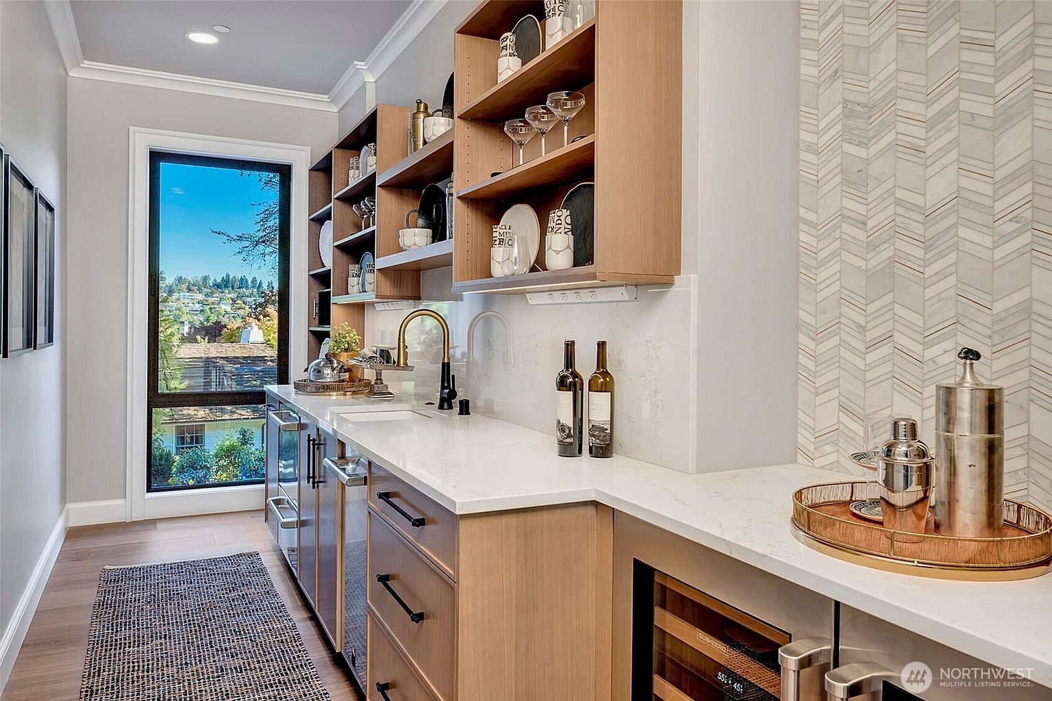 This elegant wet bar or butler's pantry features warm wood cabinetry, sleek white quartz countertops, and a sophisticated herringbone marble backsplash. The space is equipped with a stainless steel under-counter refrigerator, a wine cooler, and a modern gold faucet, all bathed in natural light from a large window overlooking a scenic neighborhood. The overall aesthetic is refined and contemporary, perfect for entertaining.