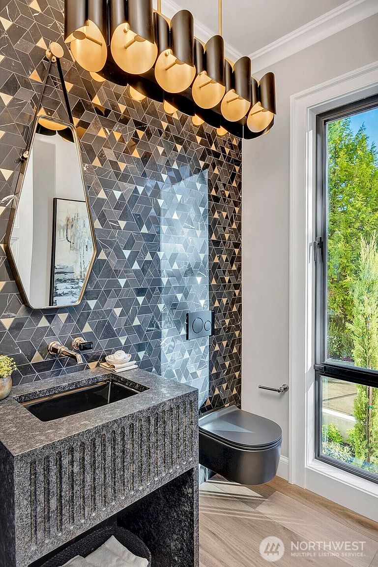 This sophisticated powder room features a striking geometric mosaic tile wall in shades of charcoal and gold, complemented by a textured stone vanity and a sleek, wall-mounted black toilet. A modern, multi-light chandelier hangs above a unique, elongated hexagonal mirror, creating a dramatic and luxurious atmosphere. The space is further enhanced by a large window that brings in natural light, highlighting the high-end finishes and contemporary design.