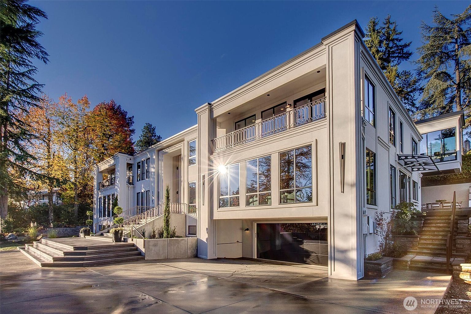 This striking modern luxury residence features a clean, white architectural facade with expansive floor-to-ceiling windows and a prominent integrated garage. The multi-level design includes elegant balconies with intricate railings and a grand staircase leading to the main entrance, all set against a backdrop of mature trees and a clear blue sky. The perspective captures the home's imposing scale and sophisticated, contemporary aesthetic, emphasizing its curb appeal and high-end construction.