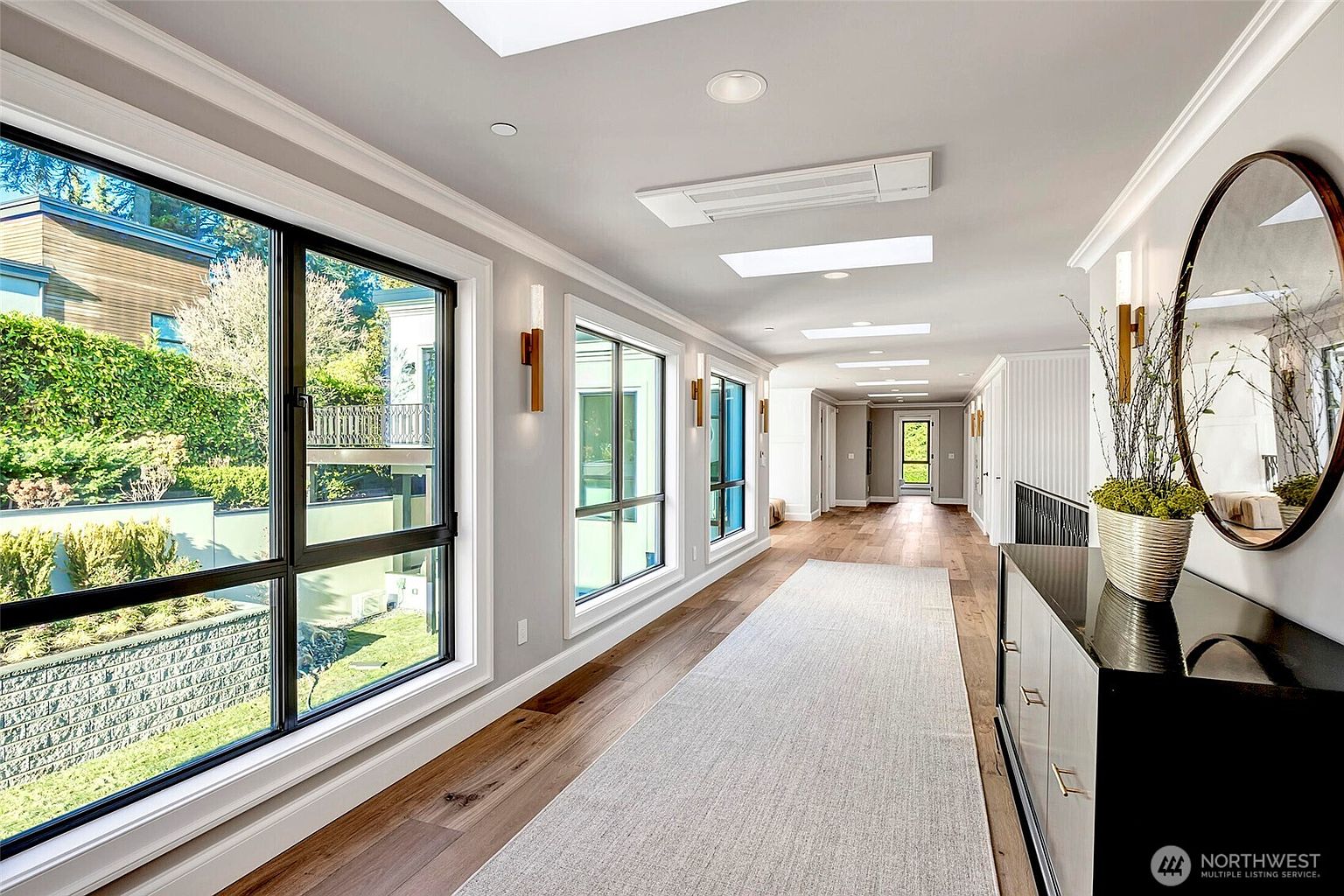 This elegant, sun-drenched hallway features rich hardwood flooring, a long runner rug, and a series of large windows that provide abundant natural light and views of the outdoors. The space is accented by modern wall sconces, a sleek console table with a circular mirror, and recessed lighting, creating a sophisticated and airy atmosphere. The perspective draws the eye down the length of the corridor toward a distant doorway, emphasizing the home's spacious and well-designed layout.