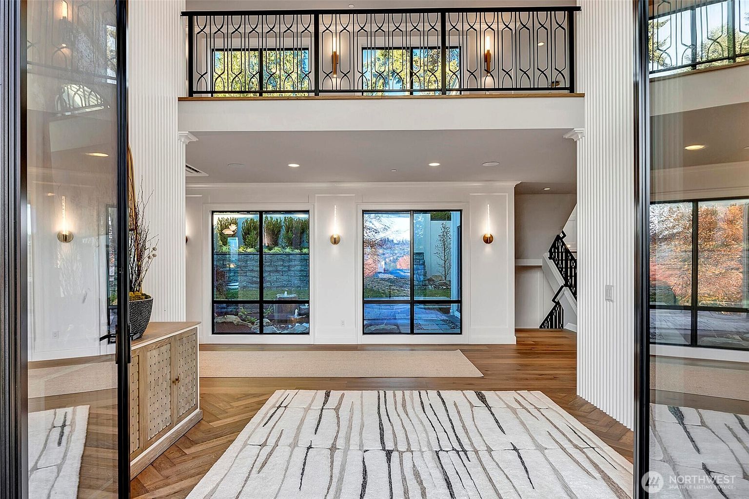 This grand, double-height entryway features a sophisticated blend of modern and classic design elements, including a striking herringbone wood floor and a textured, fluted wall finish. A sleek black metal railing overlooks the space from the second-floor landing, while large windows provide a clear view of the outdoor landscape. The perspective is captured from the threshold, emphasizing the open, airy layout and the elegant transition between the interior and exterior spaces.