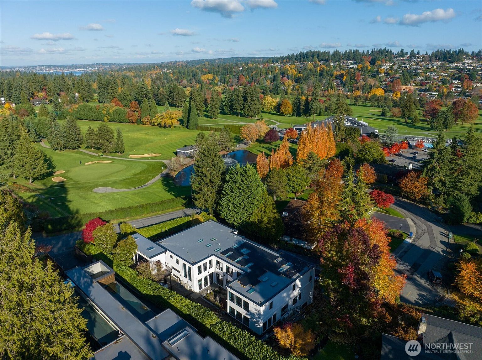 This high-angle aerial shot captures a modern, multi-level residence nestled in a lush, wooded landscape adjacent to a sprawling golf course. The home features a contemporary architectural design with a flat roof and clean lines, surrounded by vibrant autumn foliage and manicured green fairways. The perspective provides a comprehensive view of the property's prime location, highlighting its seamless integration with the surrounding natural beauty and recreational amenities.