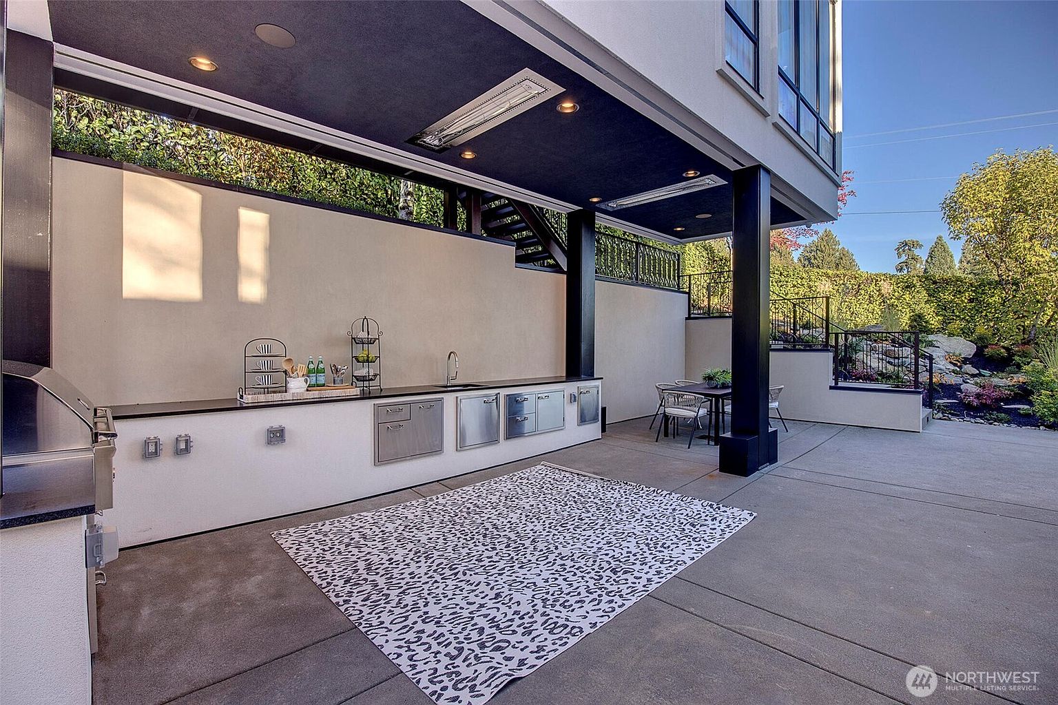This modern covered outdoor patio features a sleek built-in kitchen area with stainless steel appliances, a dark ceiling with recessed lighting, and integrated heaters. A patterned area rug sits on the concrete floor, leading toward a small dining set and a landscaped backyard with stone retaining walls. The space offers a sophisticated, contemporary design perfect for outdoor entertaining.