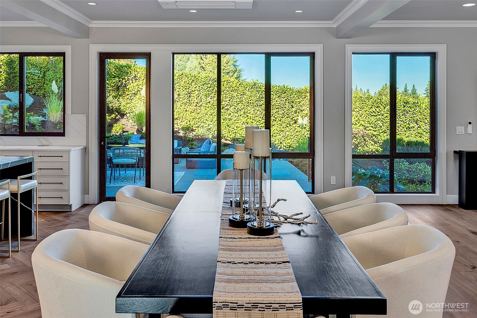 This elegant dining room features a long, dark wood table centered in the space, surrounded by modern, cream-colored upholstered chairs. Large floor-to-ceiling windows provide a seamless view of the lush green backyard and patio, flooding the room with natural light. The minimalist aesthetic is complemented by a neutral color palette and a sophisticated table runner, creating a refined and inviting atmosphere for formal dining.