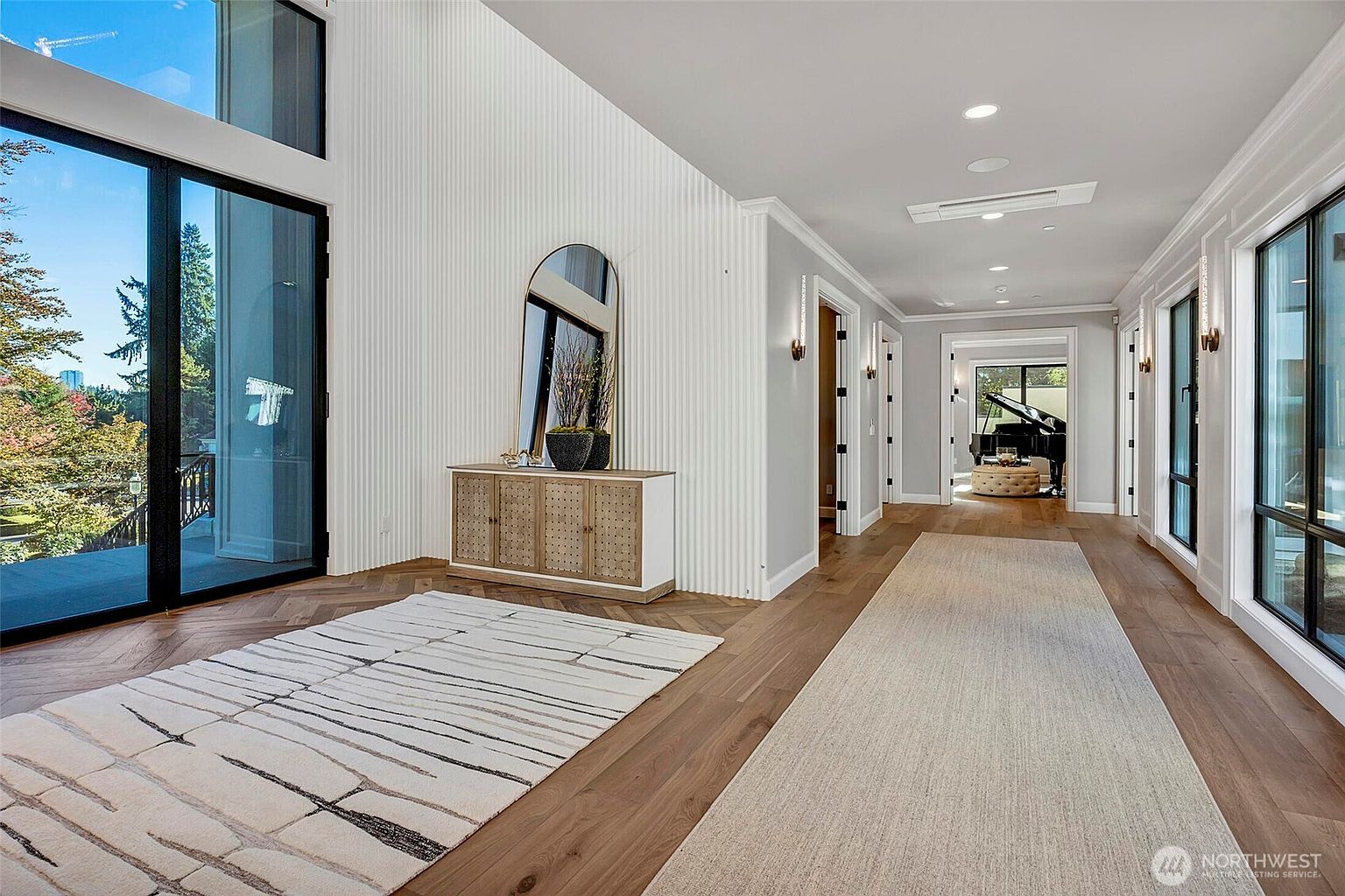 This elegant, high-ceilinged entryway and hallway features rich herringbone hardwood flooring and a striking white fluted accent wall. A modern console table with a tall arched mirror serves as a focal point, while a long runner rug leads the eye toward a bright, open space containing a grand piano. The perspective is a wide-angle shot that emphasizes the airy, sophisticated architectural design and the seamless transition between living areas.