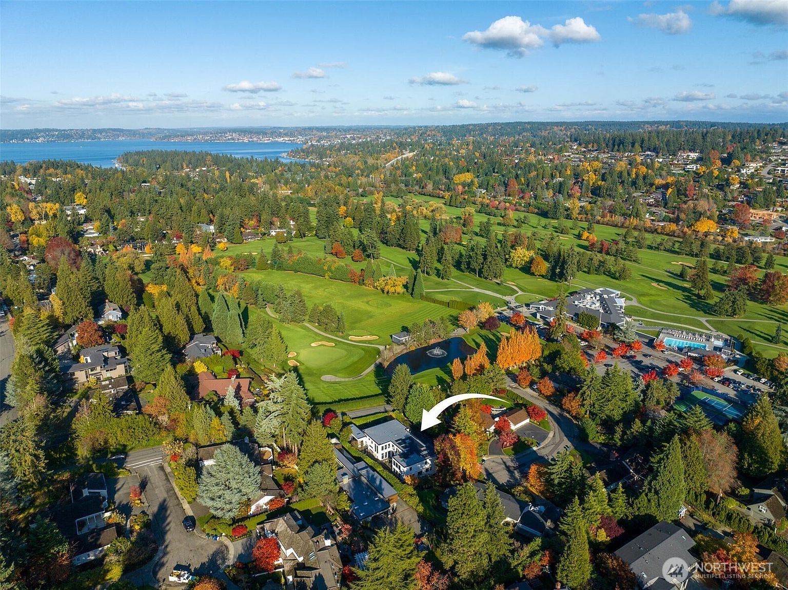 This high-angle aerial view captures a residential property nestled within a lush, tree-lined neighborhood adjacent to a sprawling golf course. The perspective highlights the home's location, showcasing its proximity to green fairways, a small pond, and the distant waterfront. The scene is bathed in natural light, emphasizing the peaceful, suburban setting and the mature landscaping surrounding the house.