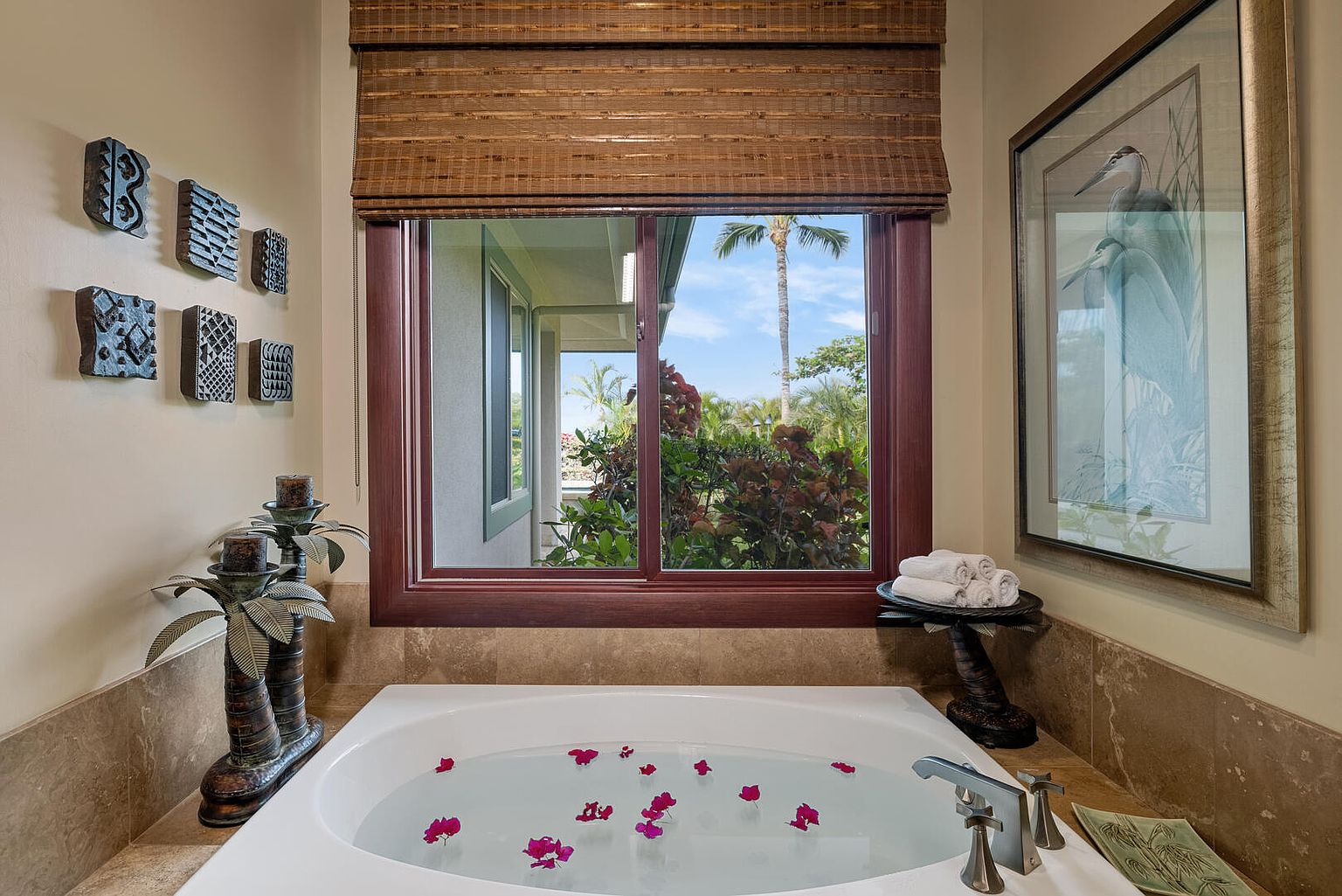 This is a luxurious primary bathroom featuring a soaking tub filled with flower petals, creating a spa-like ambiance. A large window with a woven shade offers a view of lush greenery, while artwork and decorative palm tree candle holders add to the room's elegant and tranquil atmosphere. The perspective is from slightly above the tub, looking towards the window.