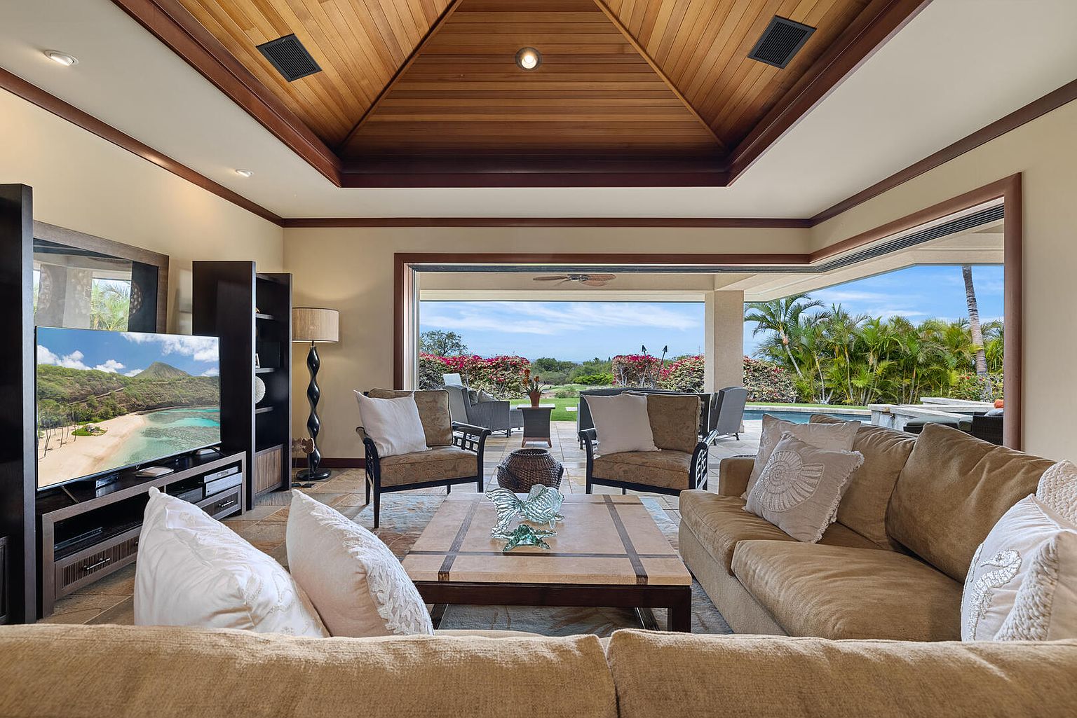 This is an interior shot of a luxurious living room featuring a coffered wood ceiling and an open floor plan that leads to an outdoor patio and pool area. The room is furnished with comfortable sofas, armchairs, and a coffee table, creating an inviting and relaxing atmosphere. The large sliding glass doors provide ample natural light and a seamless transition between indoor and outdoor living spaces.