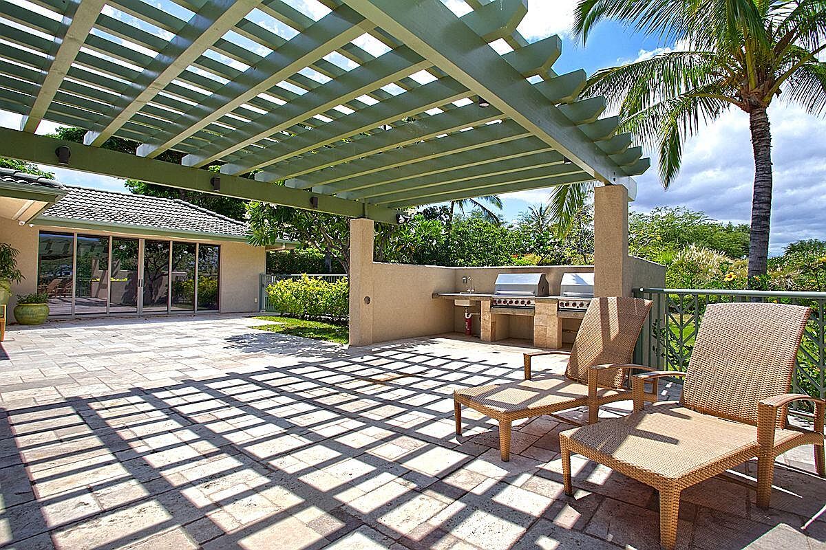 This image showcases a well-appointed outdoor patio area, featuring a pergola that casts geometric shadows across the stone-paved surface. The patio includes an outdoor kitchen with built-in grills, complemented by comfortable lounge chairs, suggesting a space designed for relaxation and entertainment. Lush greenery and a palm tree in the background enhance the tropical ambiance, making it an inviting extension of the home.