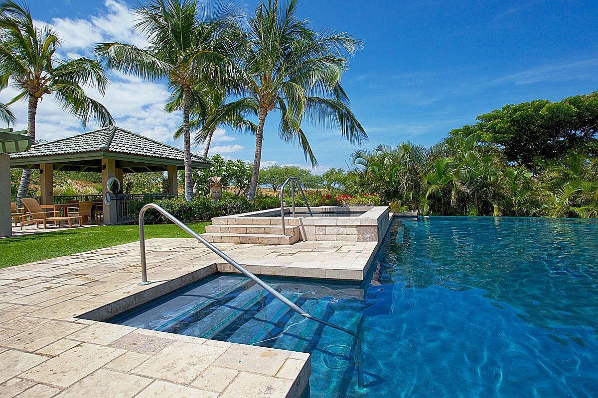 This image showcases a luxurious outdoor pool and spa area, featuring a multi-tiered design with stone steps leading into the water. Palm trees and lush tropical foliage surround the pool, creating a serene and private oasis. A covered gazebo with seating is visible in the background, suggesting an ideal space for relaxation and entertainment.