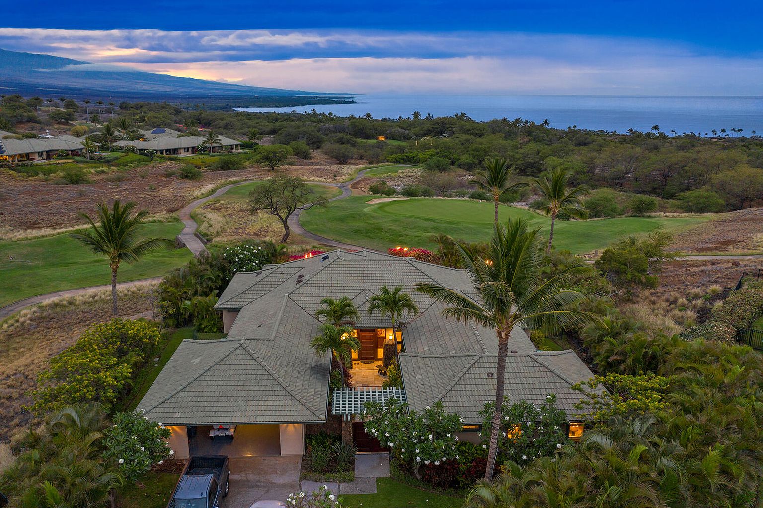 This aerial view showcases a luxurious estate nestled amidst lush greenery and a golf course, with a stunning ocean backdrop. The home features a sprawling layout with a tiled roof, complemented by mature palm trees and meticulously landscaped gardens. A glimpse of a car parked in the garage adds a touch of everyday life to this idyllic scene.