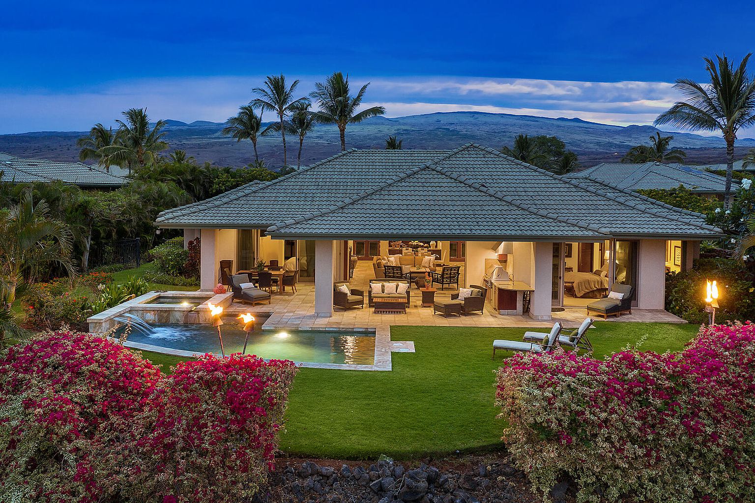 This image showcases the rear exterior of a luxurious home, featuring a seamless indoor-outdoor living space with an open patio. A pool and spa are surrounded by lush landscaping, including vibrant bougainvillea bushes. The architecture is complemented by palm trees and a mountain backdrop, creating a serene and inviting atmosphere.